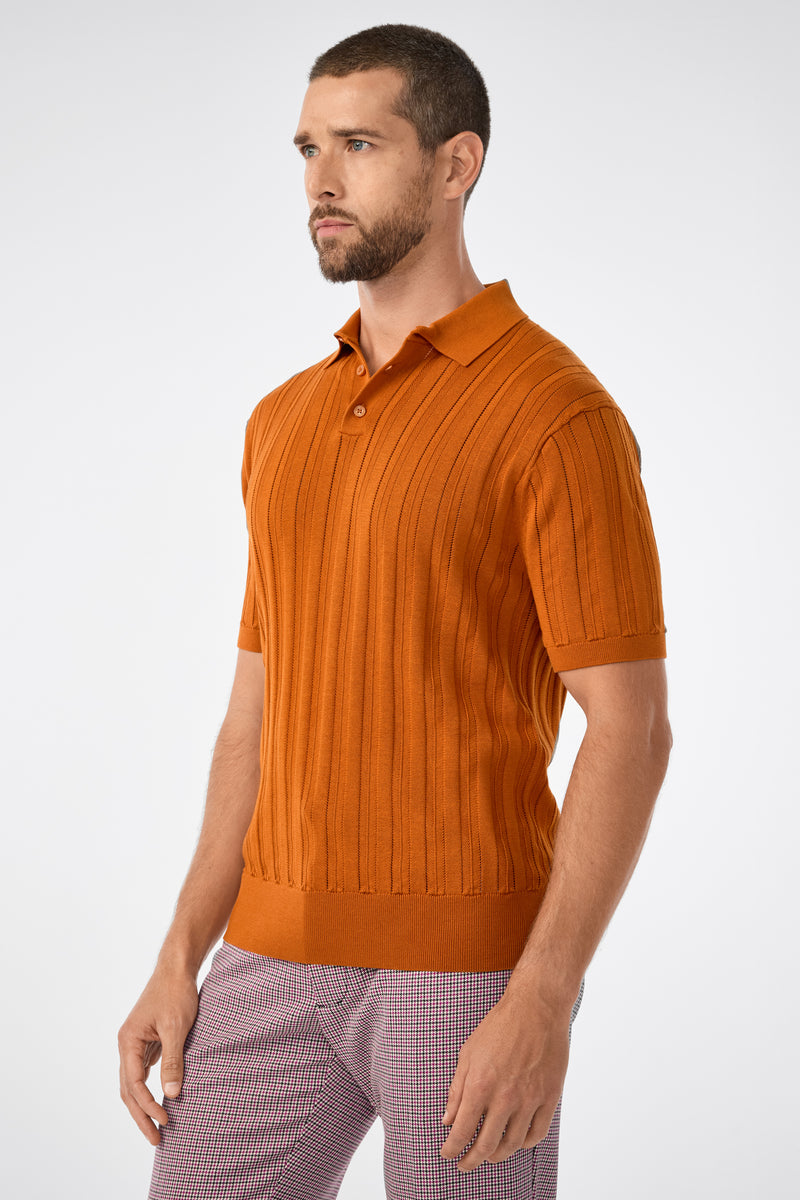 A man with short hair and a beard wears the Kensington Short-Sleeve Polo in Geranium + Amber and light checkered pants, standing against a plain white background.