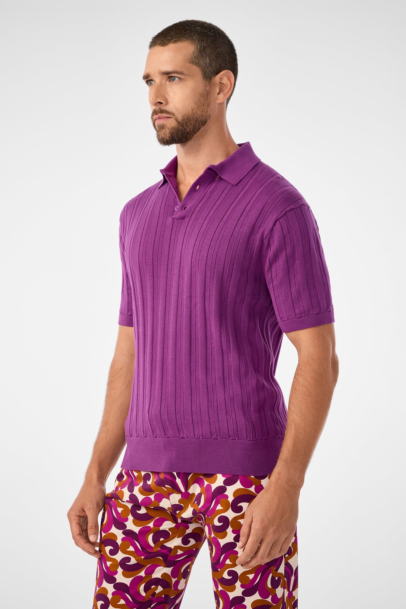 A man with short hair and a beard wears the Kensington Short-Sleeve Polo in Geranium + Amber and patterned pants with purple, gold, and white swirls, standing against a plain light background.