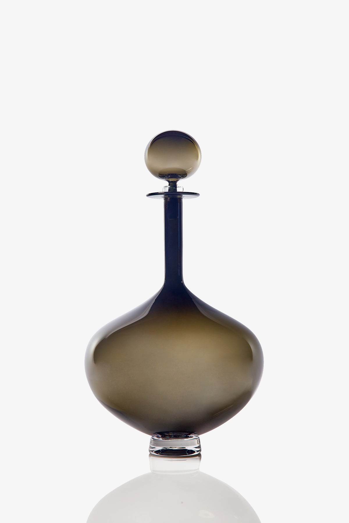 The Large Genie Bottle Decanter, crafted from smoky, dark glass with a round stopper, sits on a reflective surface against a white background.