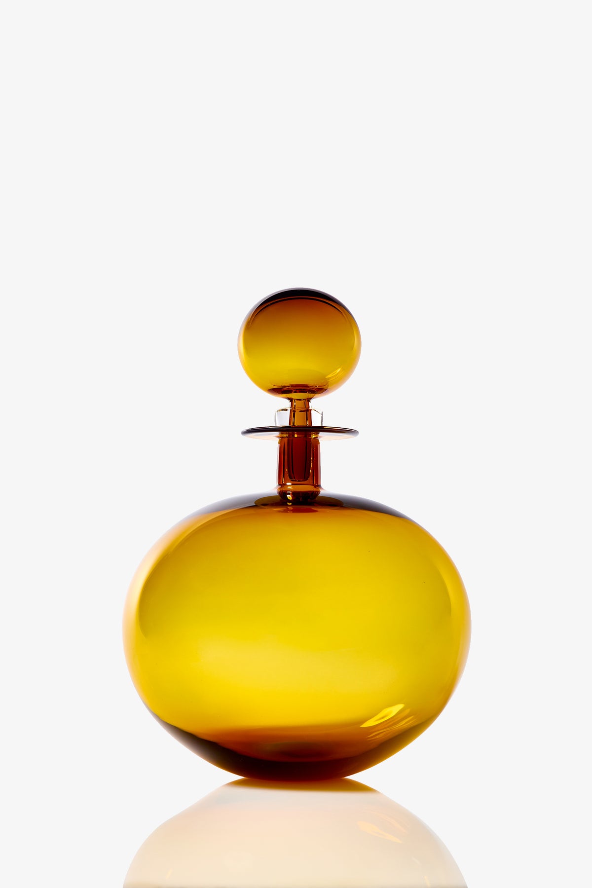 A large, low round decanter made of amber-colored glass with a matching spherical stopper, set against a plain white background. The bottle’s surface reflects light, creating subtle highlights and shadows.