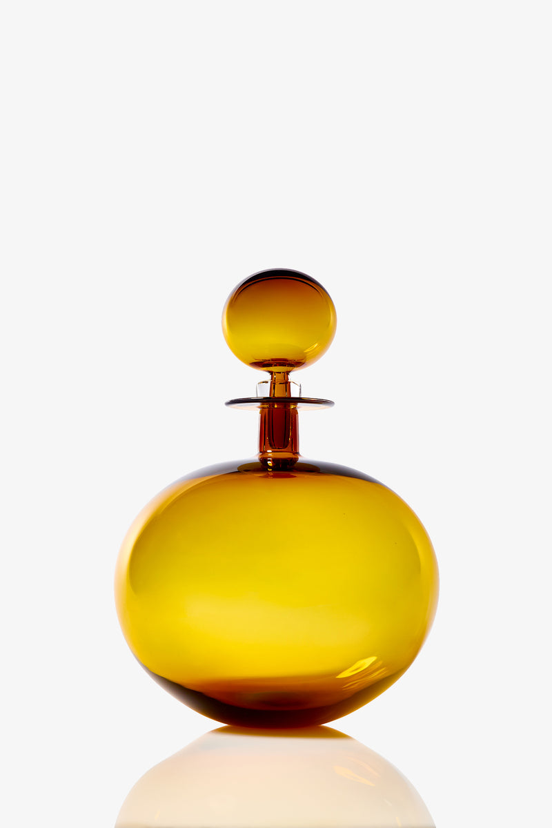 A large, low round decanter made of amber-colored glass with a matching spherical stopper, set against a plain white background. The bottle’s surface reflects light, creating subtle highlights and shadows.