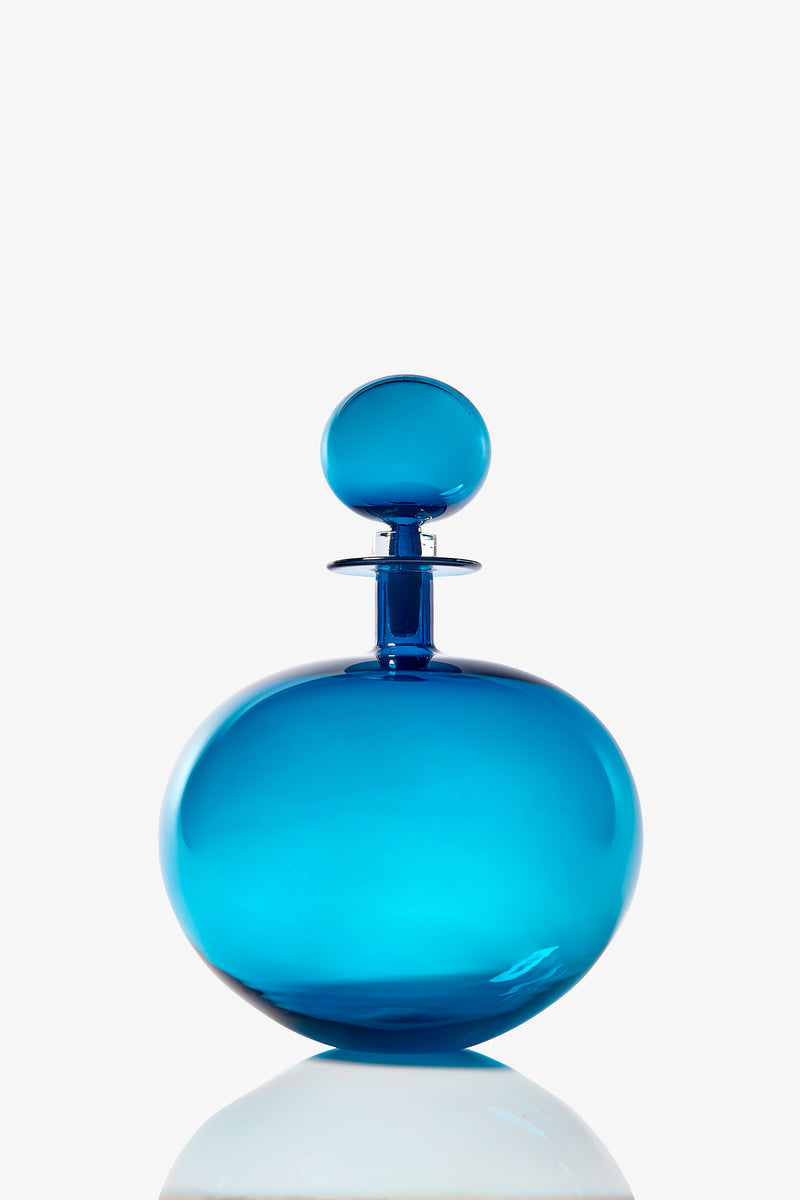 A large, round translucent blue glass decanter with a matching spherical stopper, shown against a white background and reflected on a shiny surface.