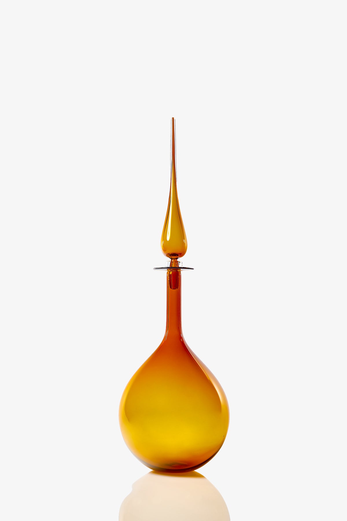 A Large Tear Drop Decanter, crafted from tall amber glass with a round base and a long pointed stopper, stands on a reflective white surface against a plain white background.