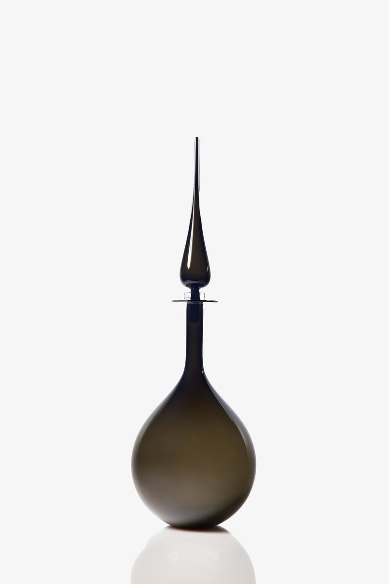 A Large Tear Drop Decanter with a dark, opaque glass body, wide base, and long pointed stopper stands upright against a white background, casting a faint reflection below.