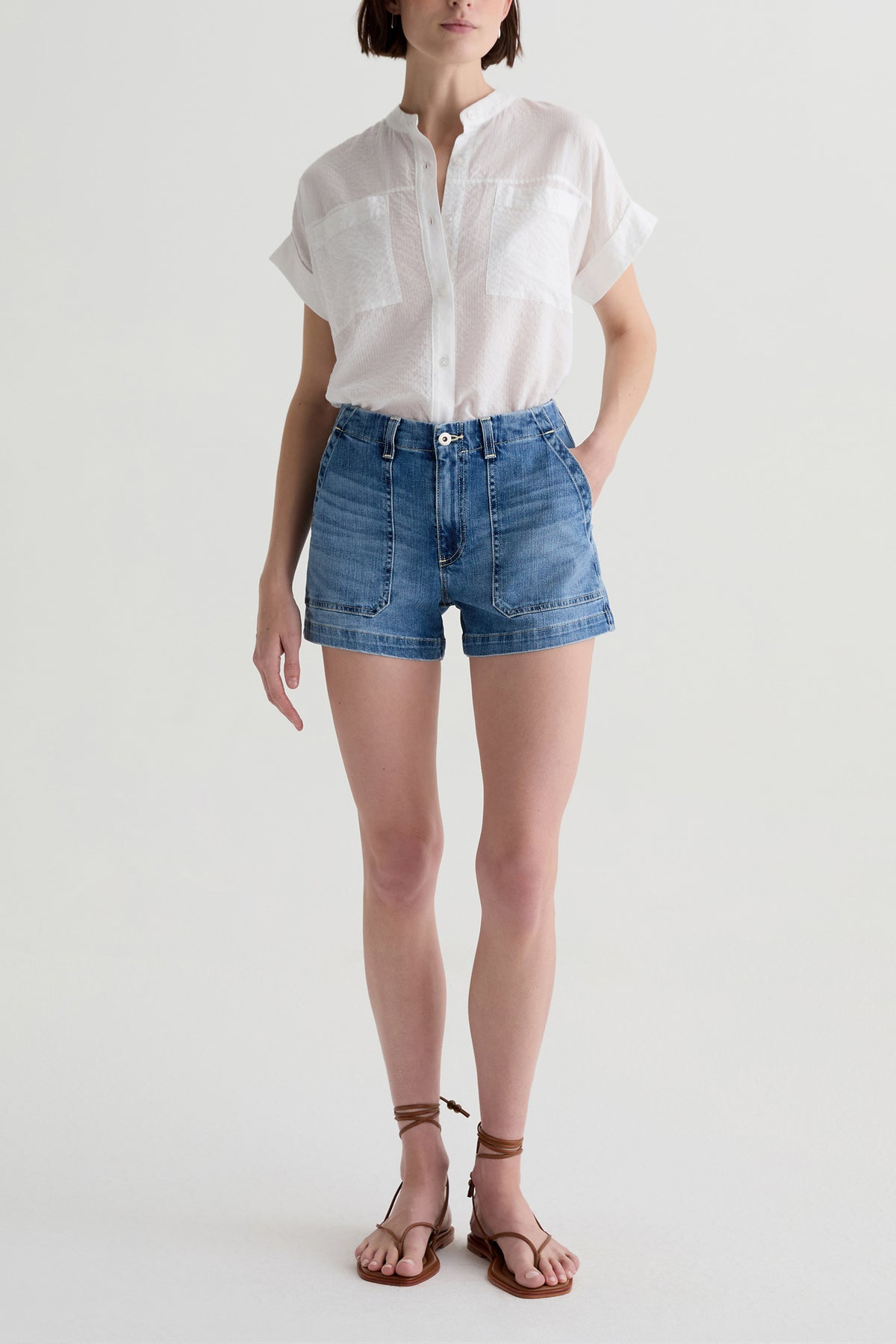 A person wears a white short-sleeve button-up shirt, AG Analeigh High-Rise Shorts, and brown strappy sandals while standing against a plain light background.