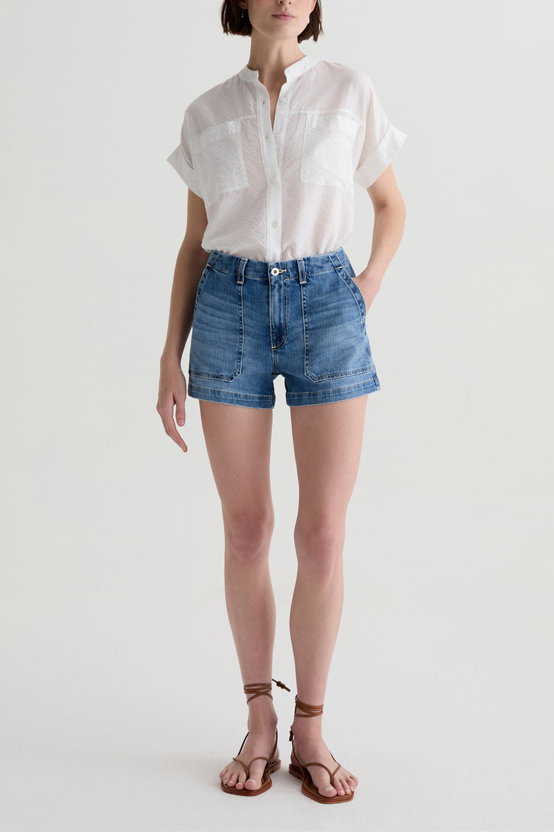 A person wears a white short-sleeve button-up shirt, AG Analeigh High-Rise Shorts, and brown strappy sandals while standing against a plain light background.