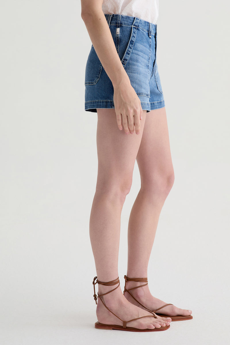 A person stands sideways against a plain light background wearing the AG Analeigh High-Rise Short in blue denim, paired with a white top and brown strappy flat sandals.