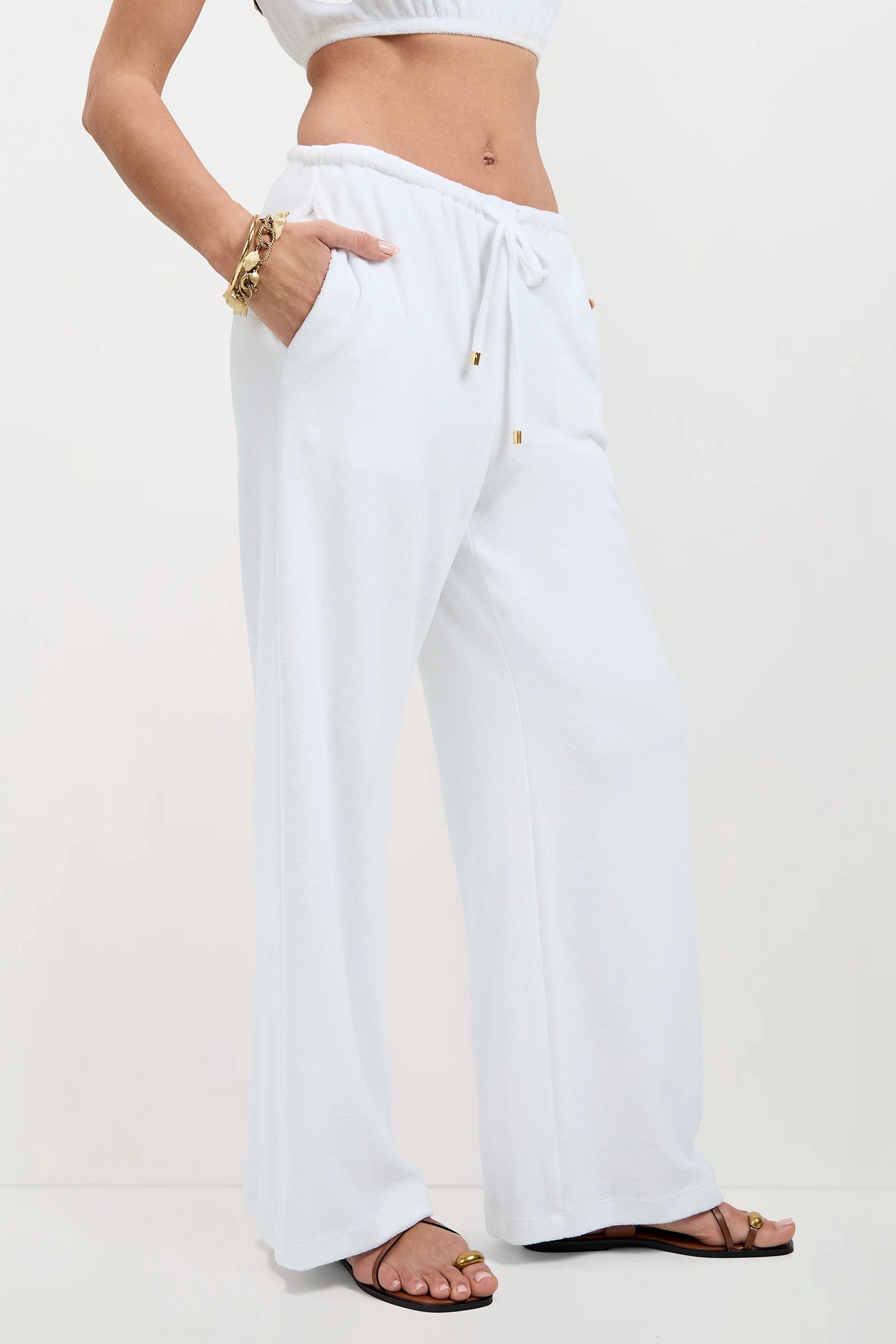 A woman wears the Linsk Wide-Leg Pant - Poolside Terry in white, paired with a cropped white top, gold bracelets, and flat sandals. She stands with one hand in her pocket; only her body from mid-torso down is shown against a plain backdrop.
