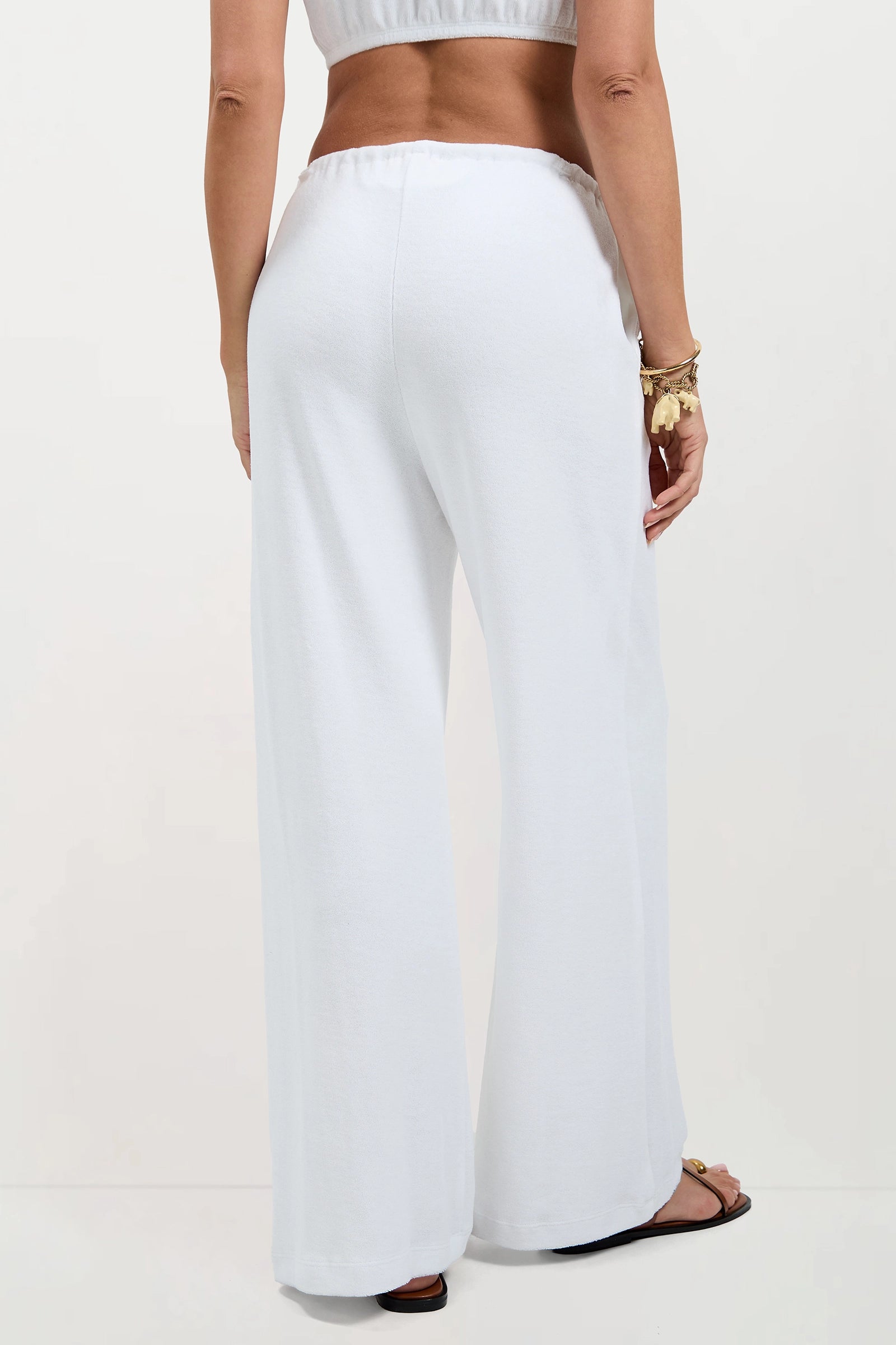 A person is shown from the back wearing the Linsk Wide-Leg Pant - Poolside Terry paired with a matching white top, sandals, and a gold charm bracelet on their left wrist.