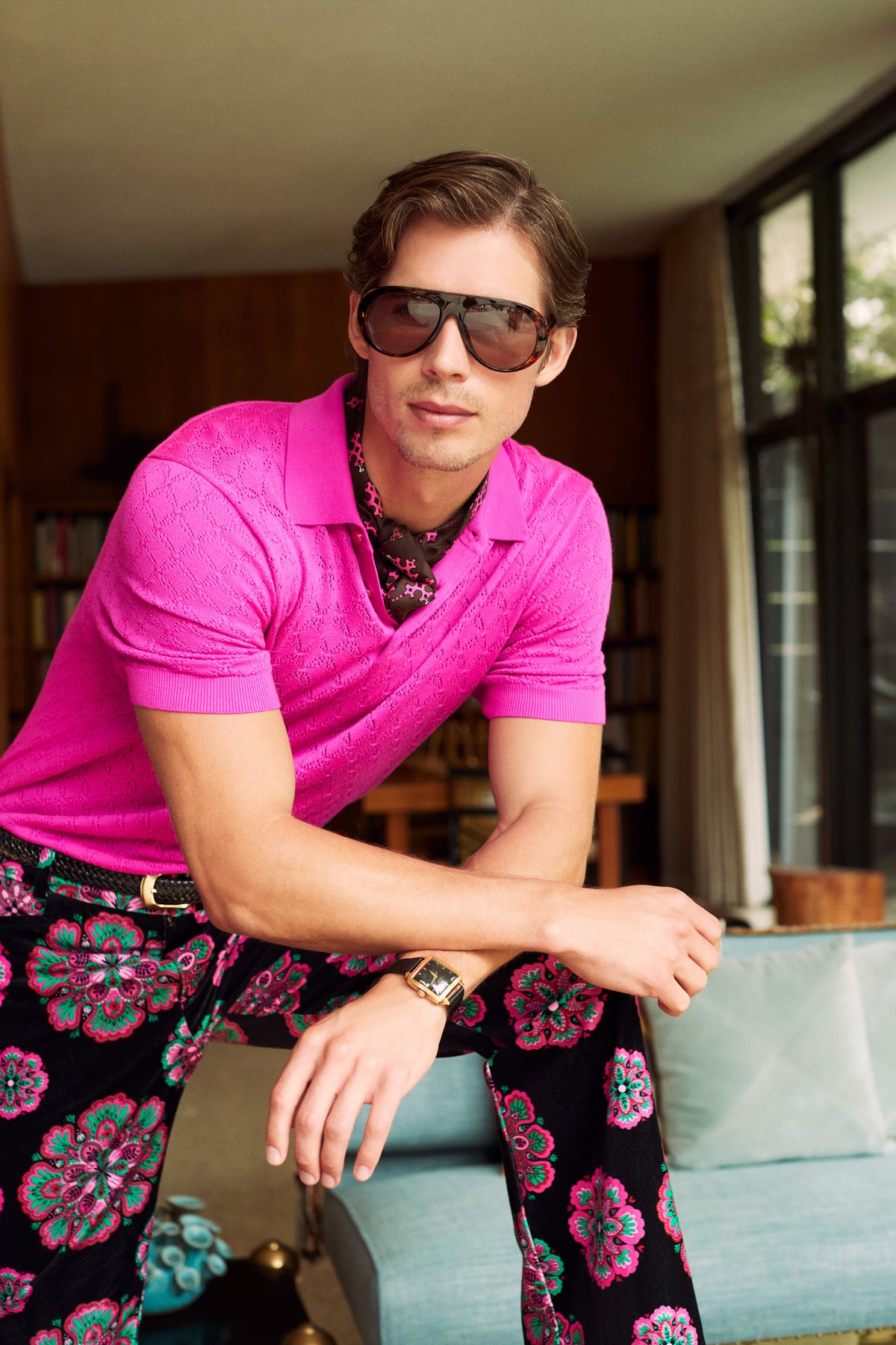 A man wearing a short-sleeved hot pink Waylen Polo and patterned floral trousers leans forward while wearing large sunglasses.