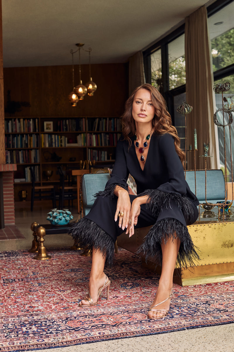 A woman sits wearing the black Anina Top and matching cropped trousers, both trimmed with dark feathers, paired with gold strappy heels and a large multi-colored statement necklace.