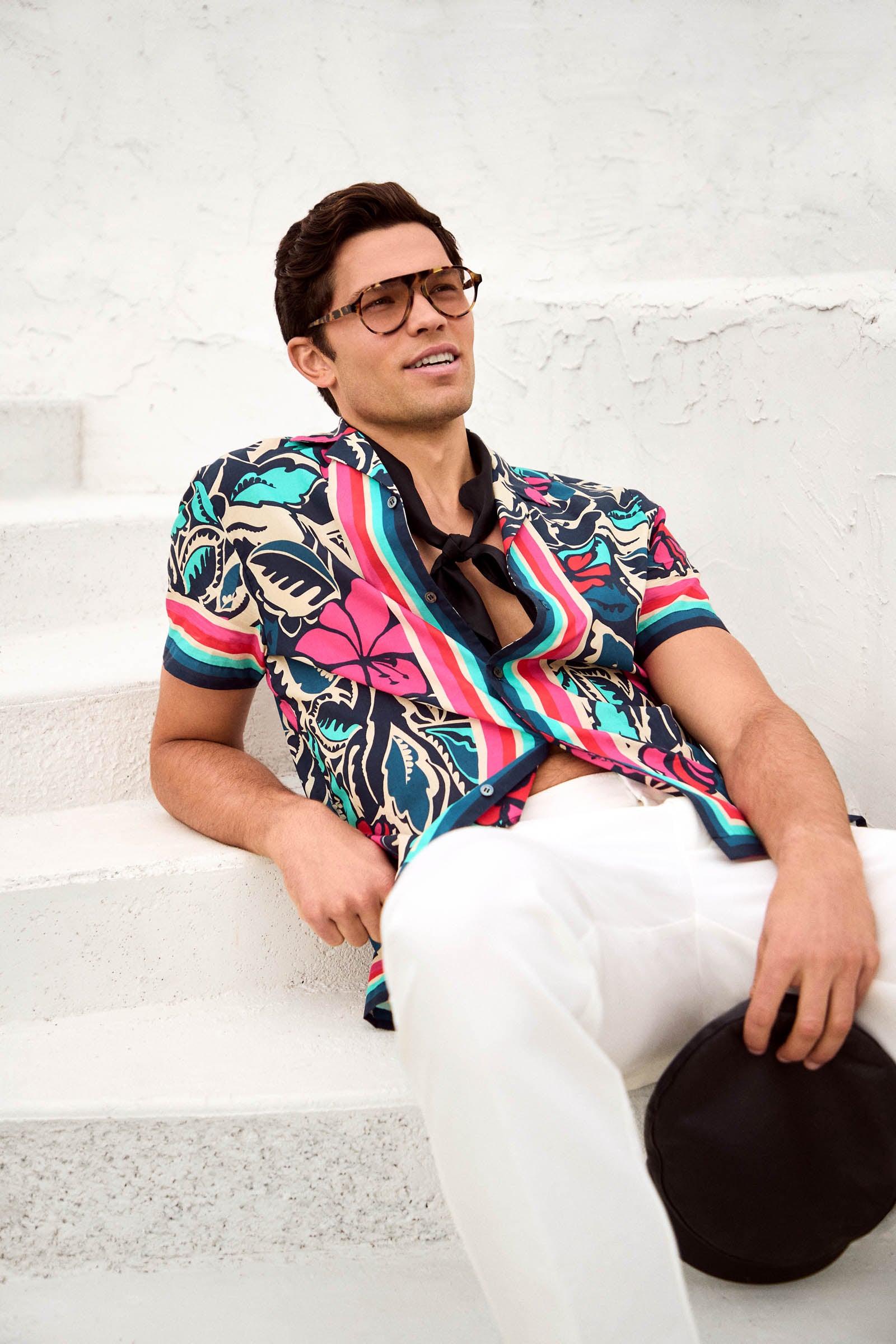 A man wearing the Josue Short-Sleeve Shirt - Copacabana and white pants relaxes on white steps, holding a black hat in a bright outdoor setting.