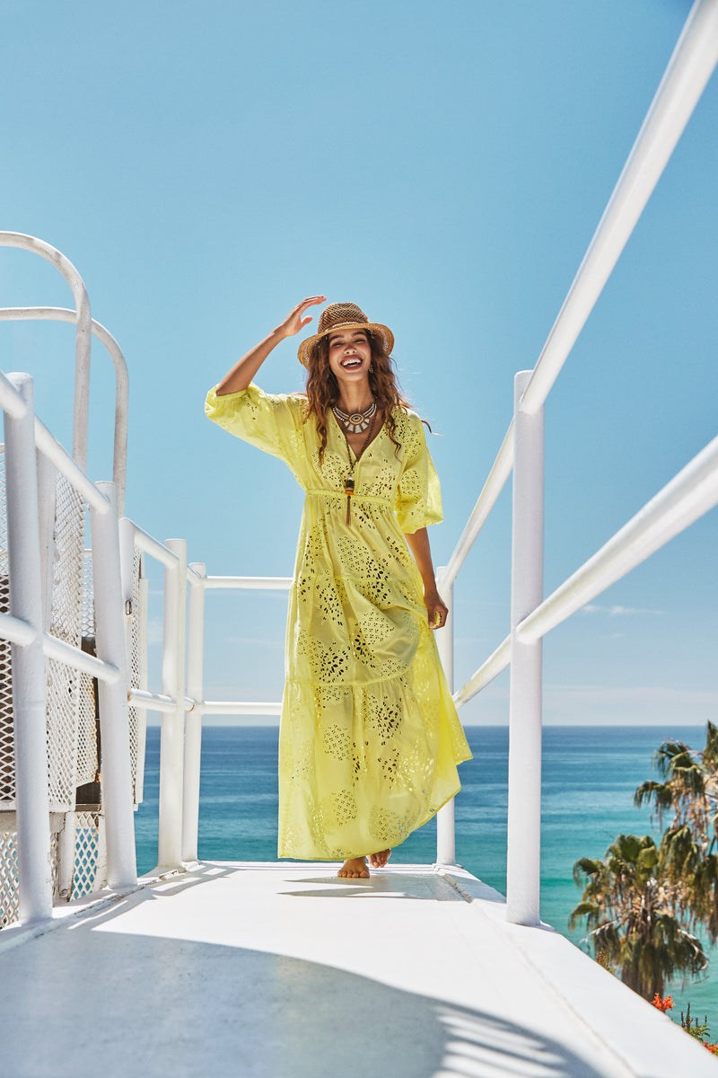 A woman smiles barefoot on a sunlit walkway by the ocean, wearing the bright yellow Arco Iris Dress and a straw hat, with palm trees and a clear blue sky in the background.