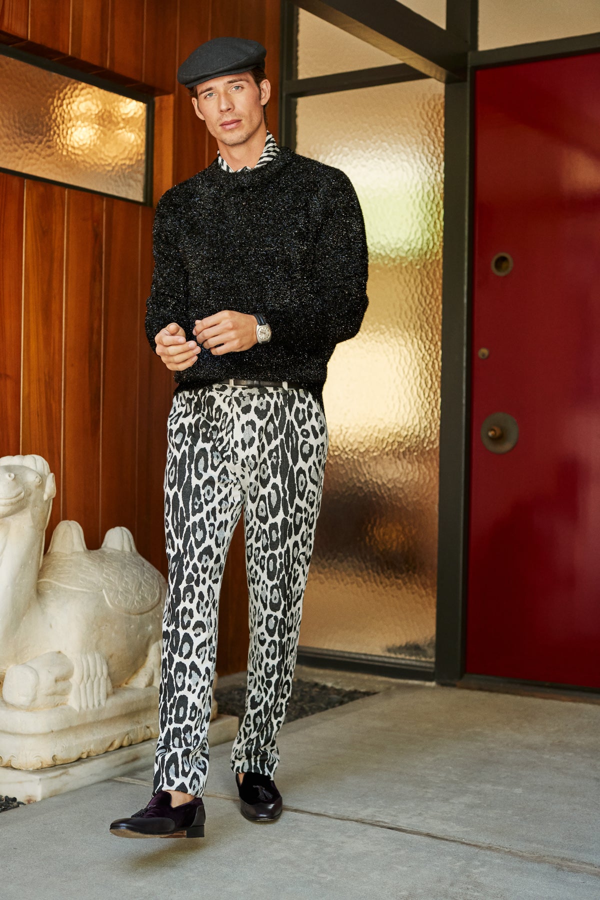 A man wears a dark flat cap, a black sweater, and black-and-white leopard print Clyde Slim Trousers.