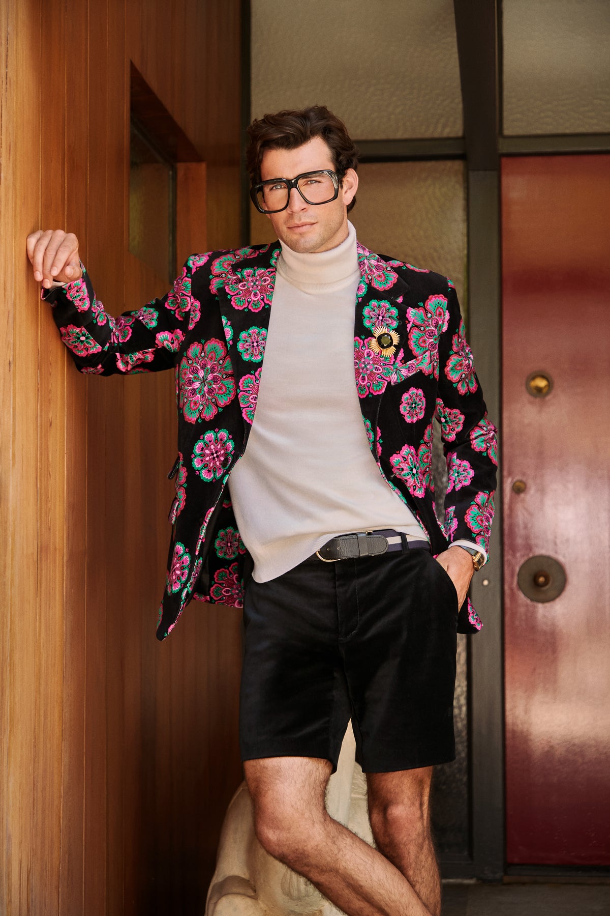 A man wears a Doheny Blazer in a black, pink, and teal floral pattern over a beige turtleneck and black shorts.