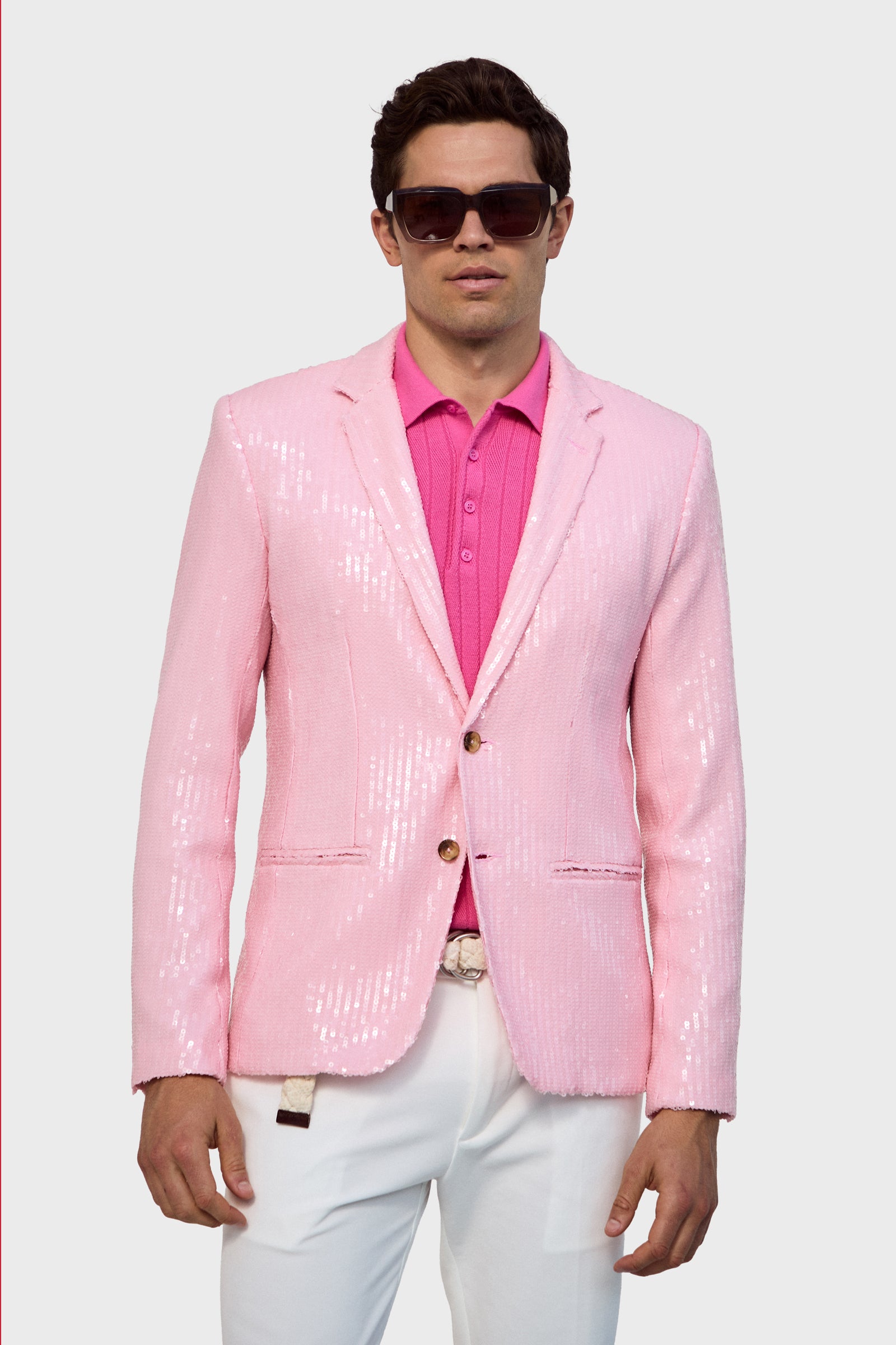 A man wearing the Bowie Sequin Blazer in shiny light pink over a bright pink shirt, white pants, and large dark sunglasses poses against a plain light background.