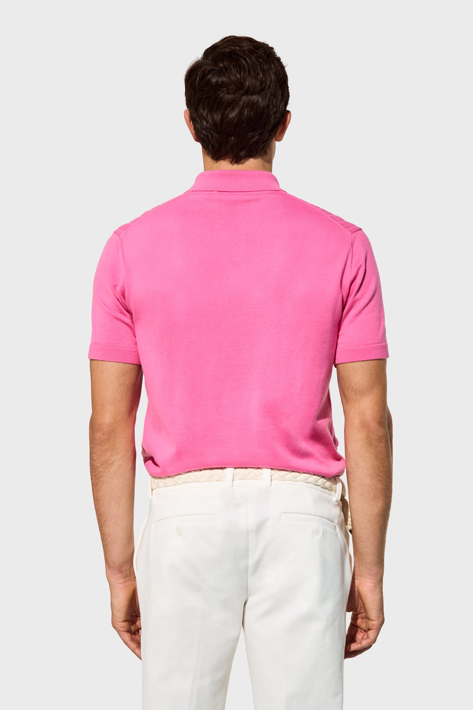 A man seen from behind wears the Alvarez Short-Sleeve Polo in bright pink with white trousers, standing against a plain light gray background.
