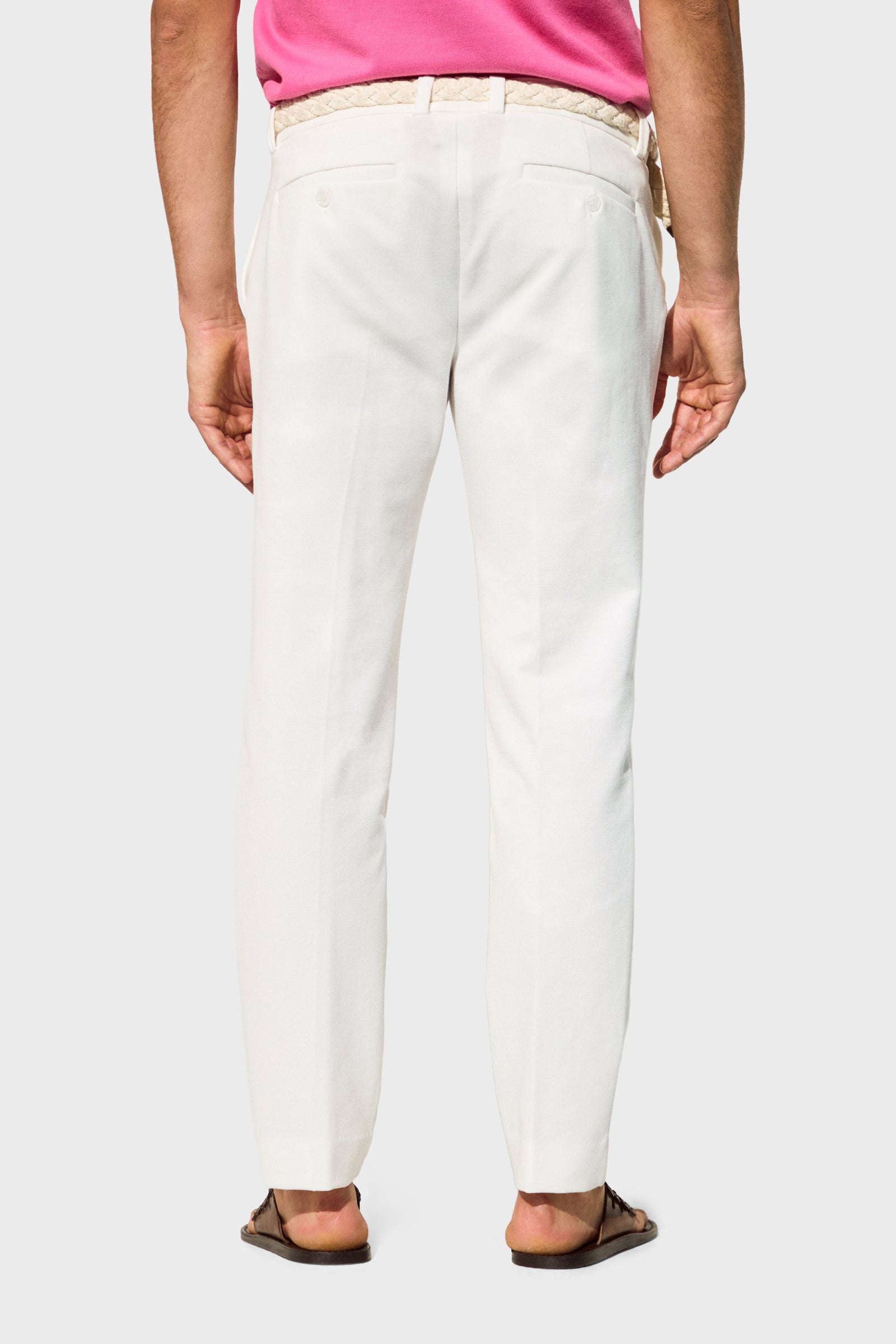 A person shown from the back against a plain, light background wears Clyde Slim Trouser - Stretch Piqué in white, styled with a braided belt, pink shirt, and brown sandals.