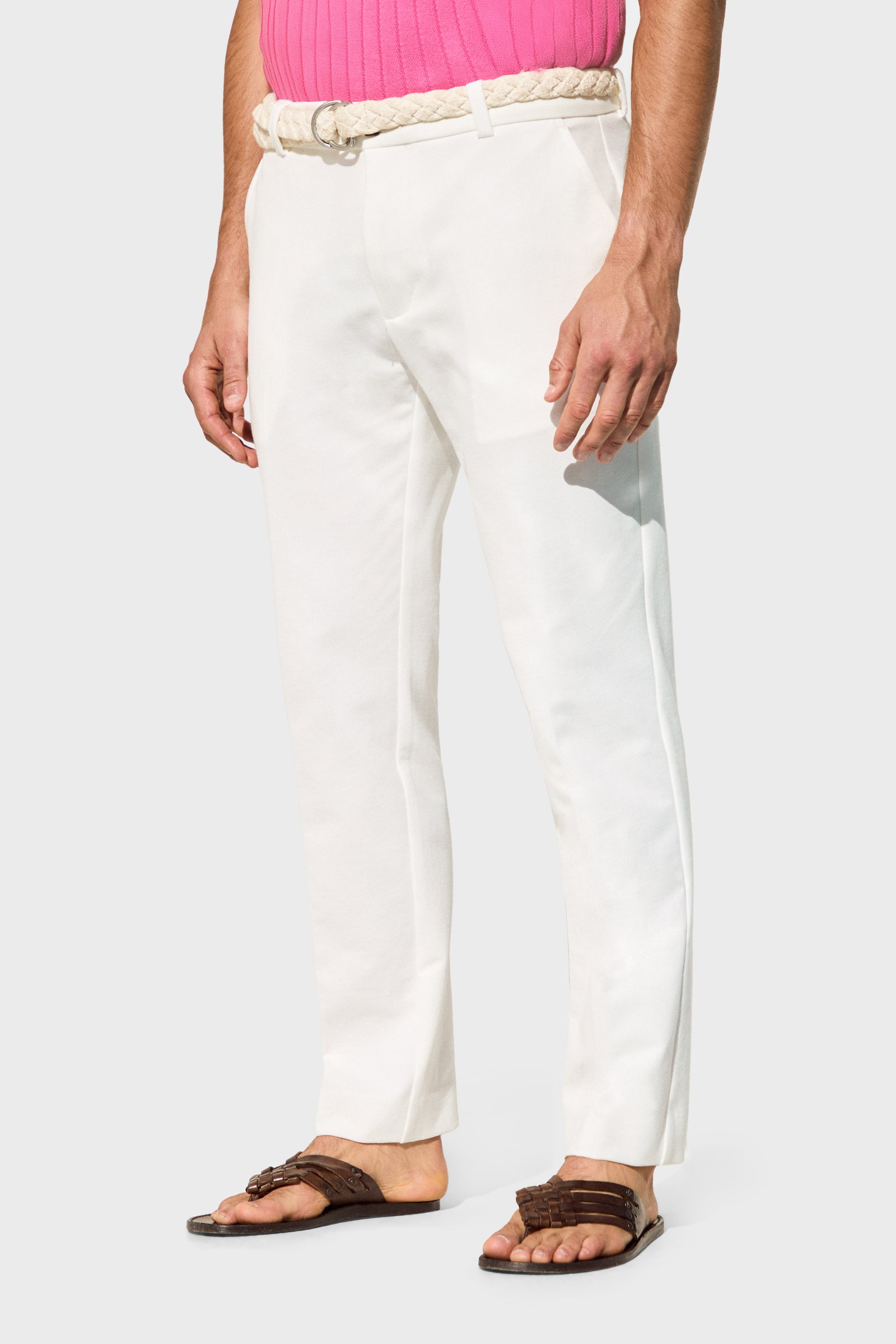 A person wears the Clyde Slim Trouser - Stretch Piqué in white with a braided belt, pink top, and brown sandals against a plain light gray background, hands relaxed at their sides.