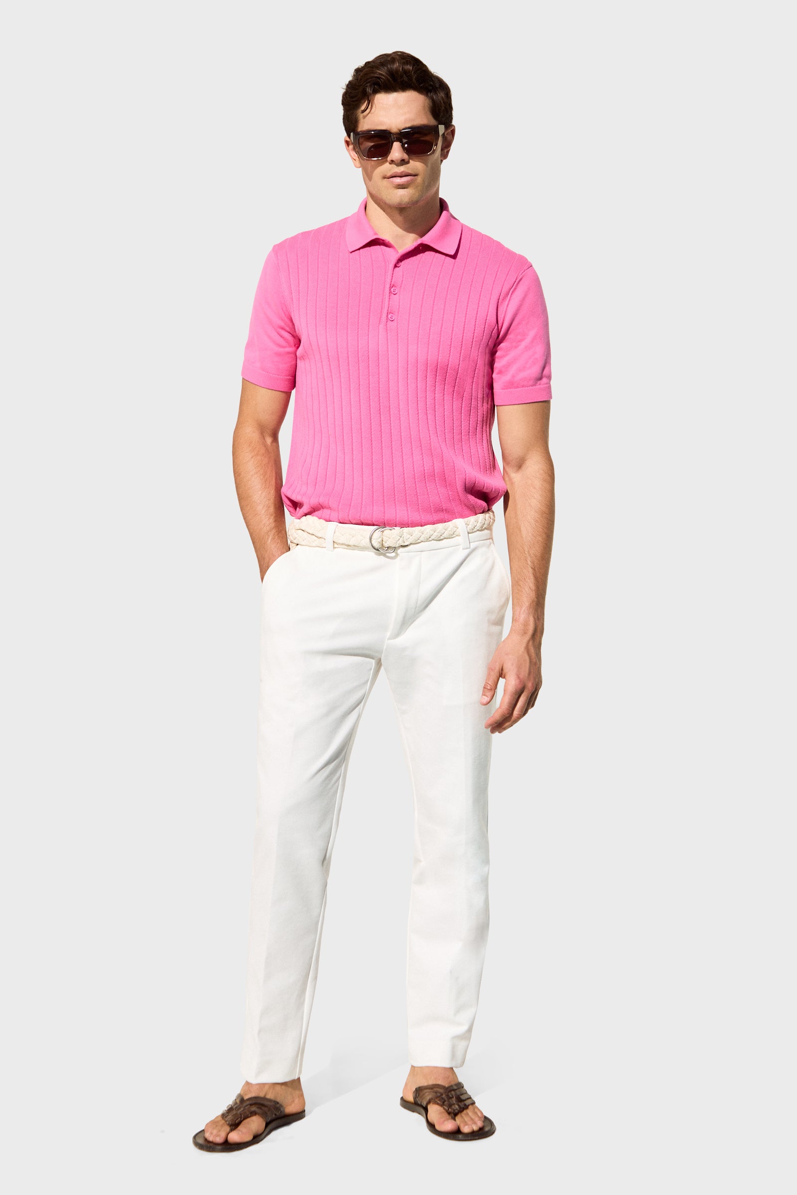 A man wearing sunglasses, a bright pink short-sleeve polo, and the Clyde Slim Trouser - Stretch Piqué stands against a plain light gray background.