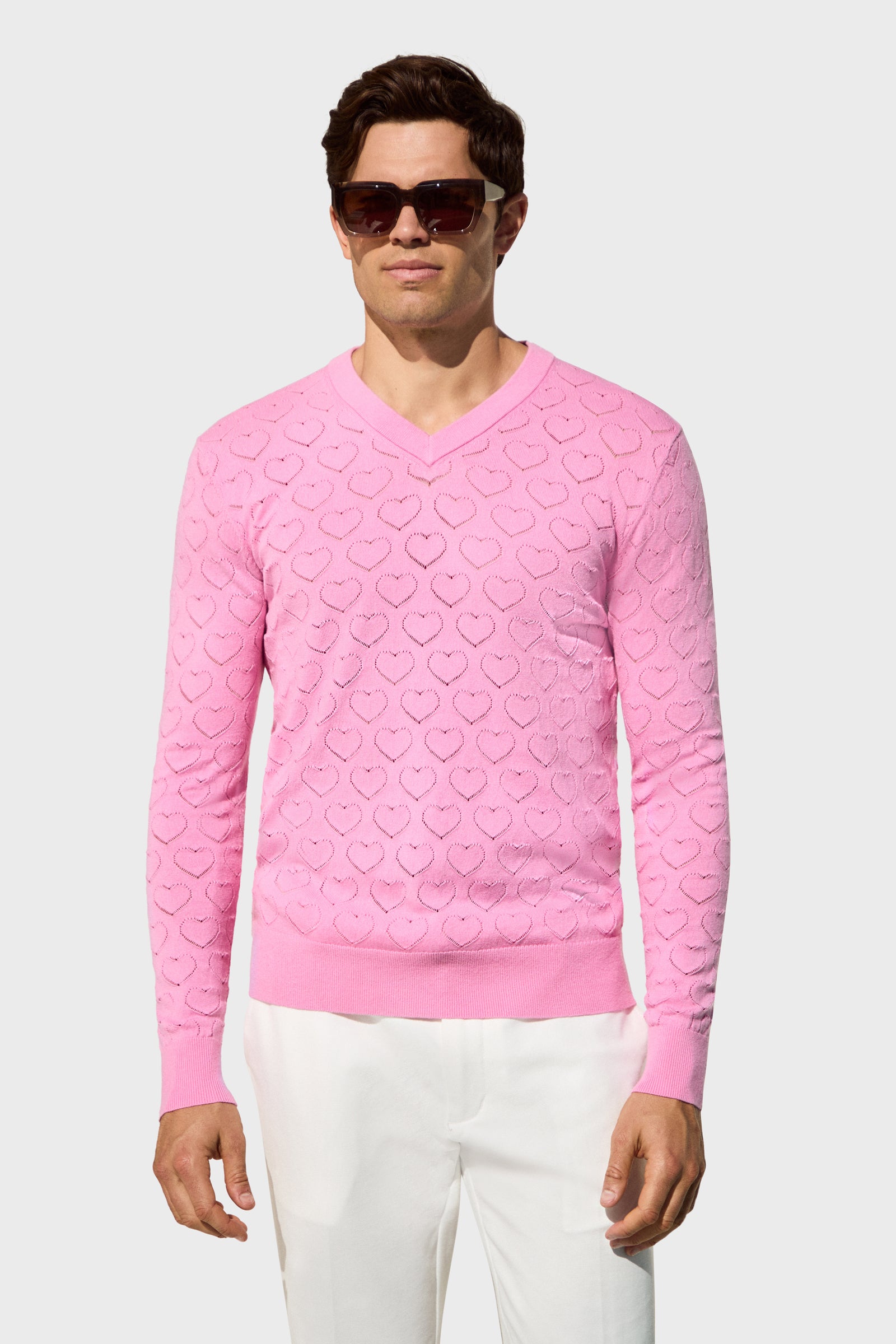 A man wears the Eros Heart Pointelle sweater in light pink with a heart pattern, paired with white pants and sunglasses, standing against a plain light gray background.