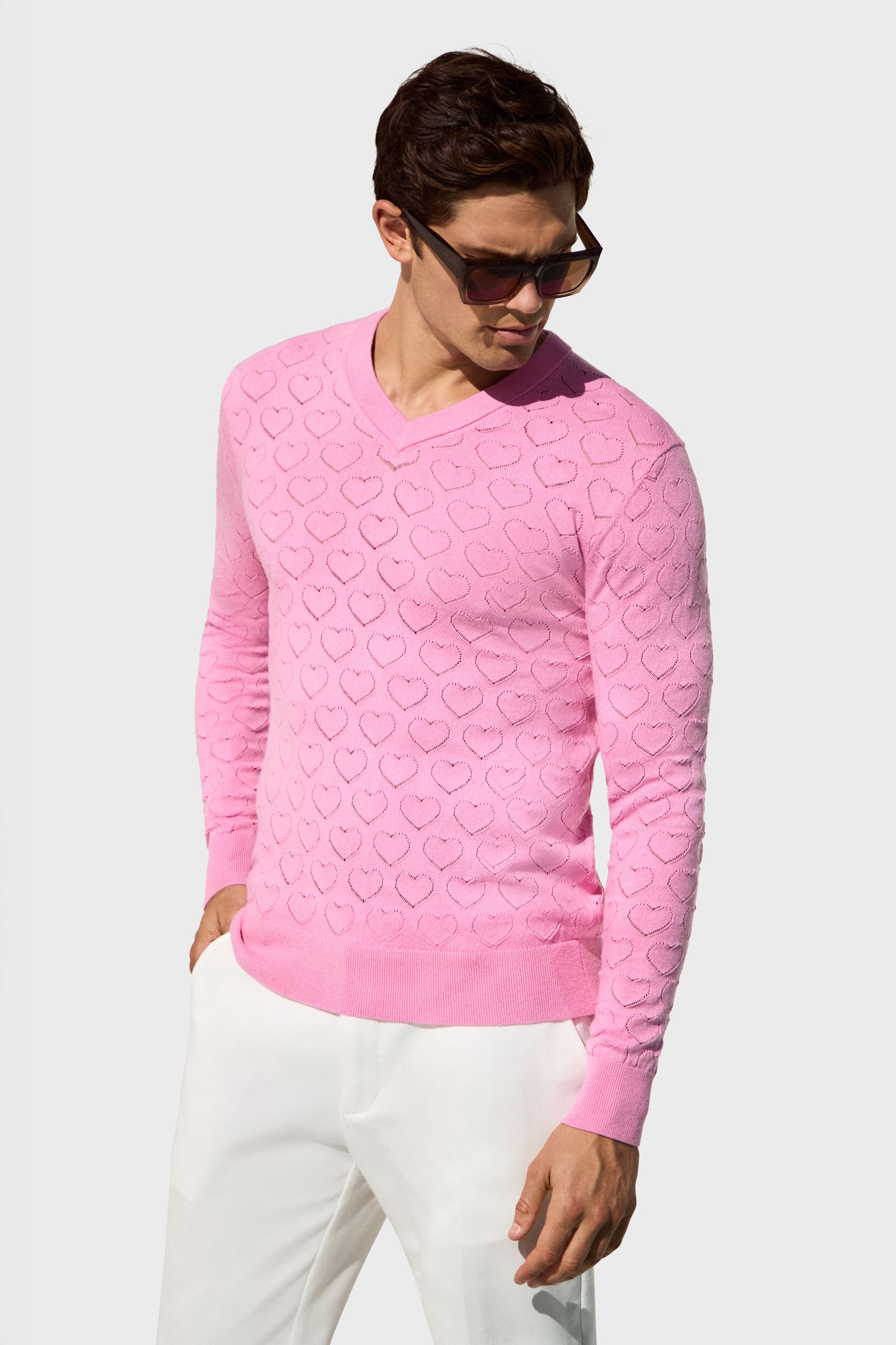A man models the Eros Heart Pointelle, a light pink heart-patterned sweater, paired with white pants and sunglasses, posing with his left hand in his pocket against a plain light gray background.