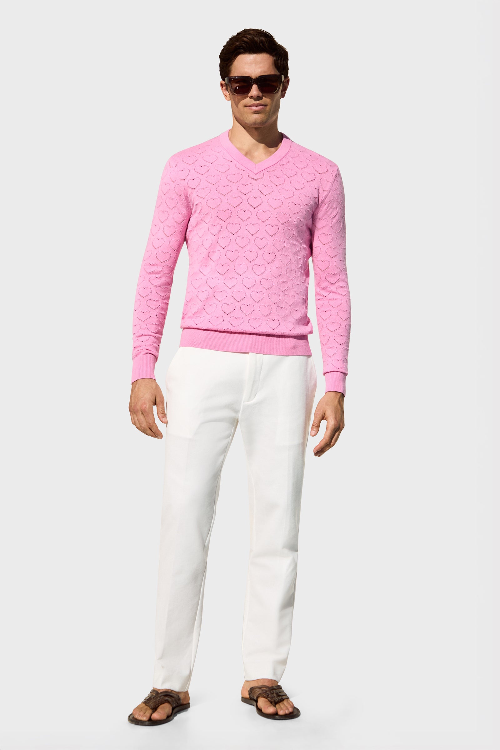 A man wearing the Eros Heart Pointelle—a light pink heart-patterned long-sleeve sweater—sunglasses, white pants, and brown sandals stands facing forward against a plain light background.