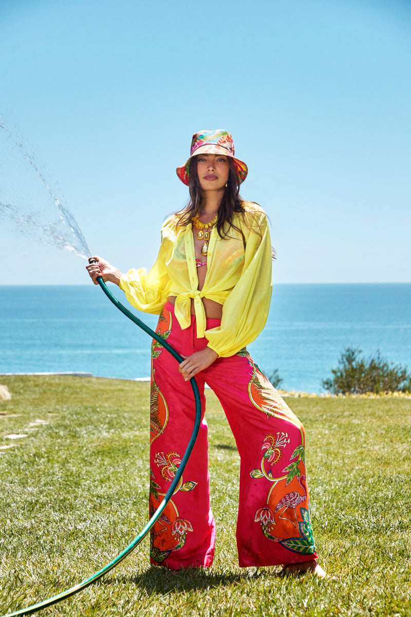 A woman waters grass on a sunny lawn by the ocean, wearing red floral Lanay Pants, a yellow blouse, and a matching hat.