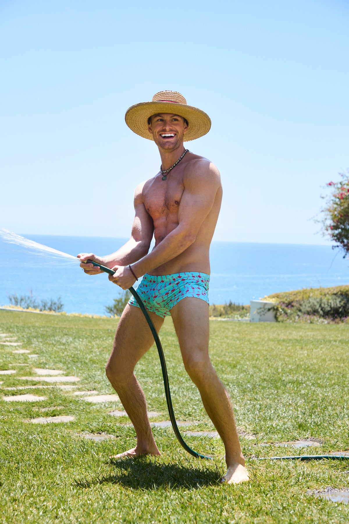 A smiling man in Sorrento Swim Trunk and a straw hat stands barefoot on grass, spraying water with a garden hose against an ocean and blue sky backdrop.