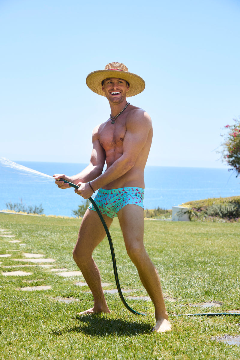 A smiling man in Sorrento Swim Trunk and a straw hat stands barefoot on grass, spraying water with a garden hose against an ocean and blue sky backdrop.