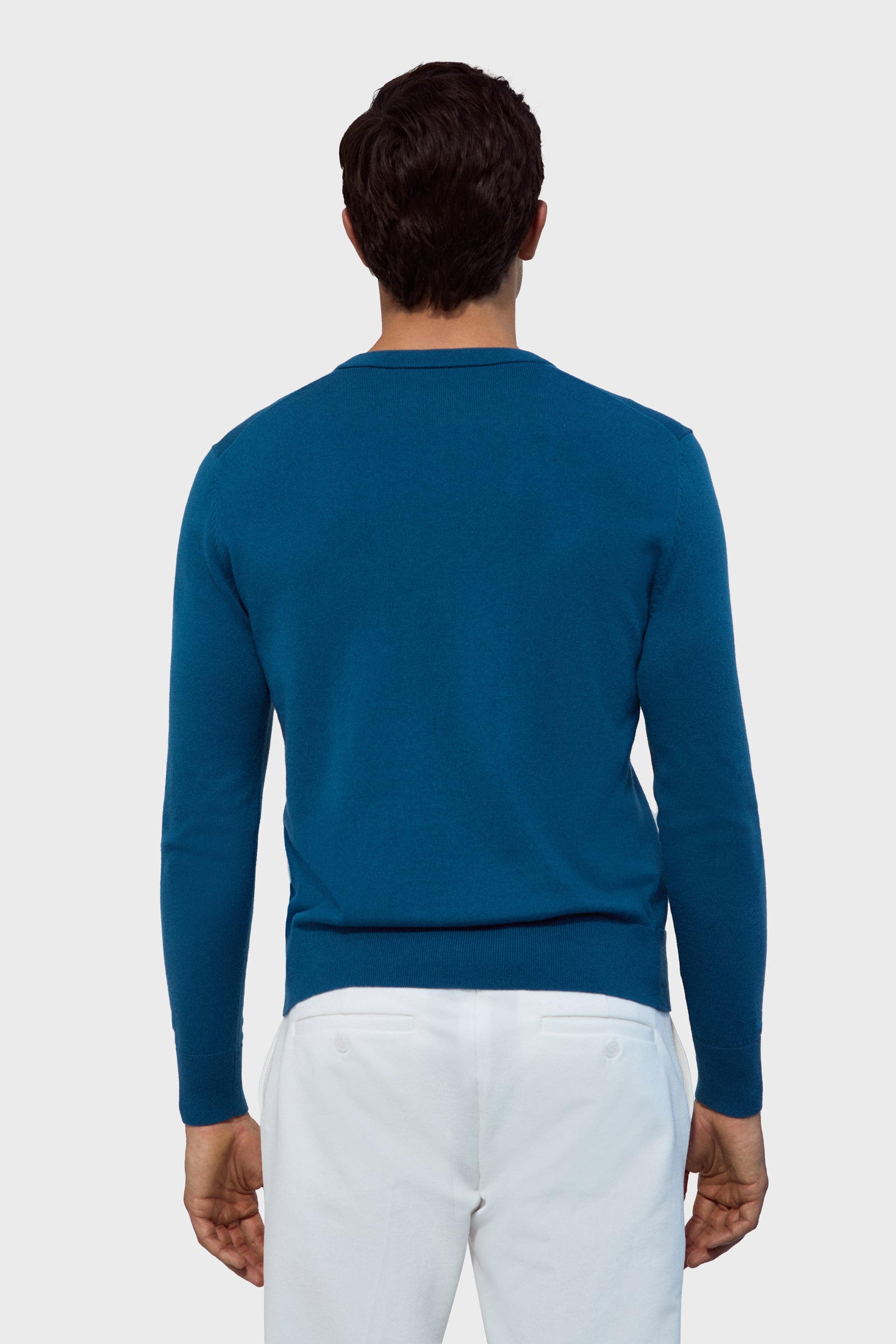 A man stands with his back to the camera wearing the Copacabana V-Neck Sweater in blue and white pants, set against a plain light gray background.