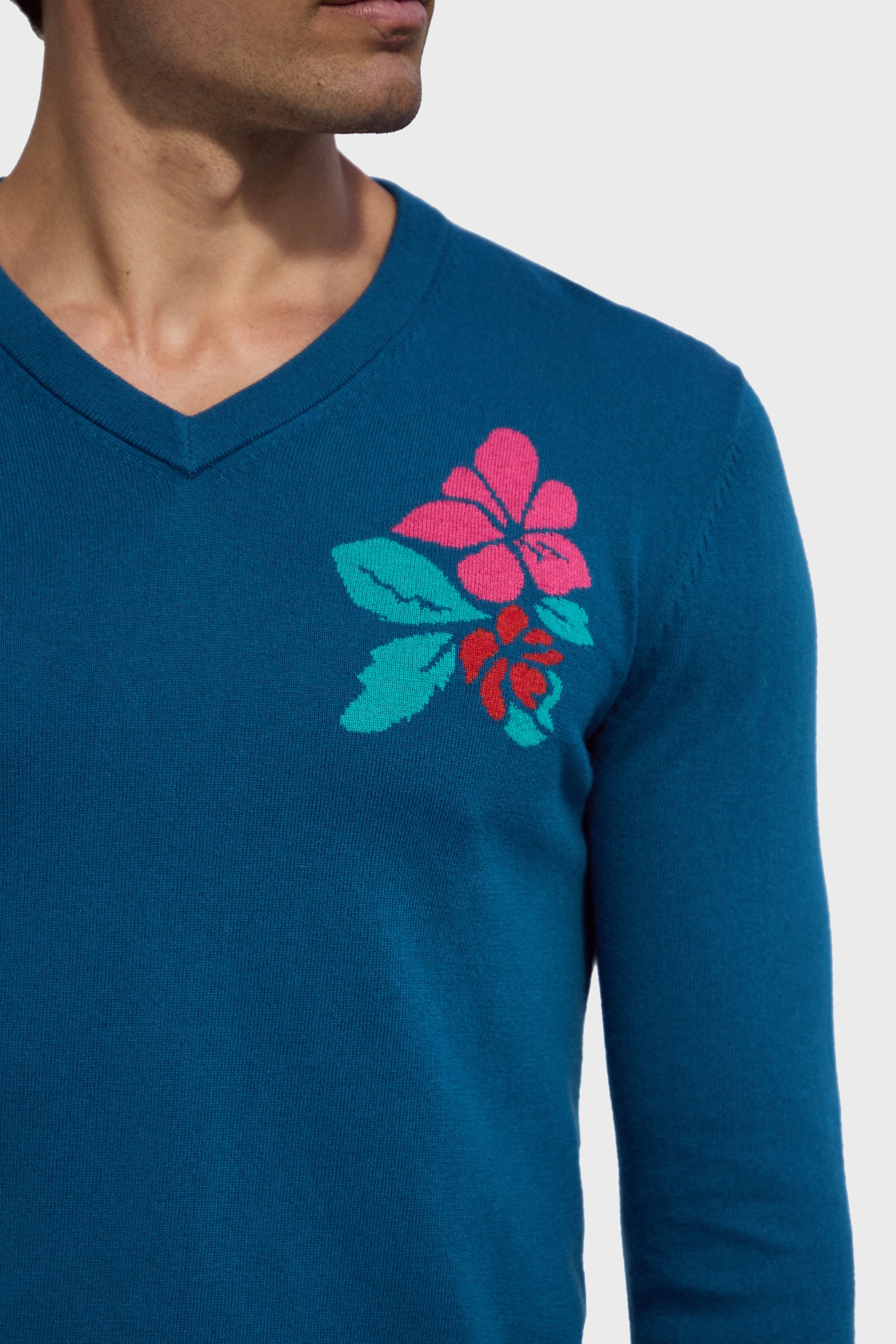 A man wears the Copacabana V-Neck Sweater, featuring a blue base with bright pink and red floral designs and green leaves on the left chest, set against a plain white background.