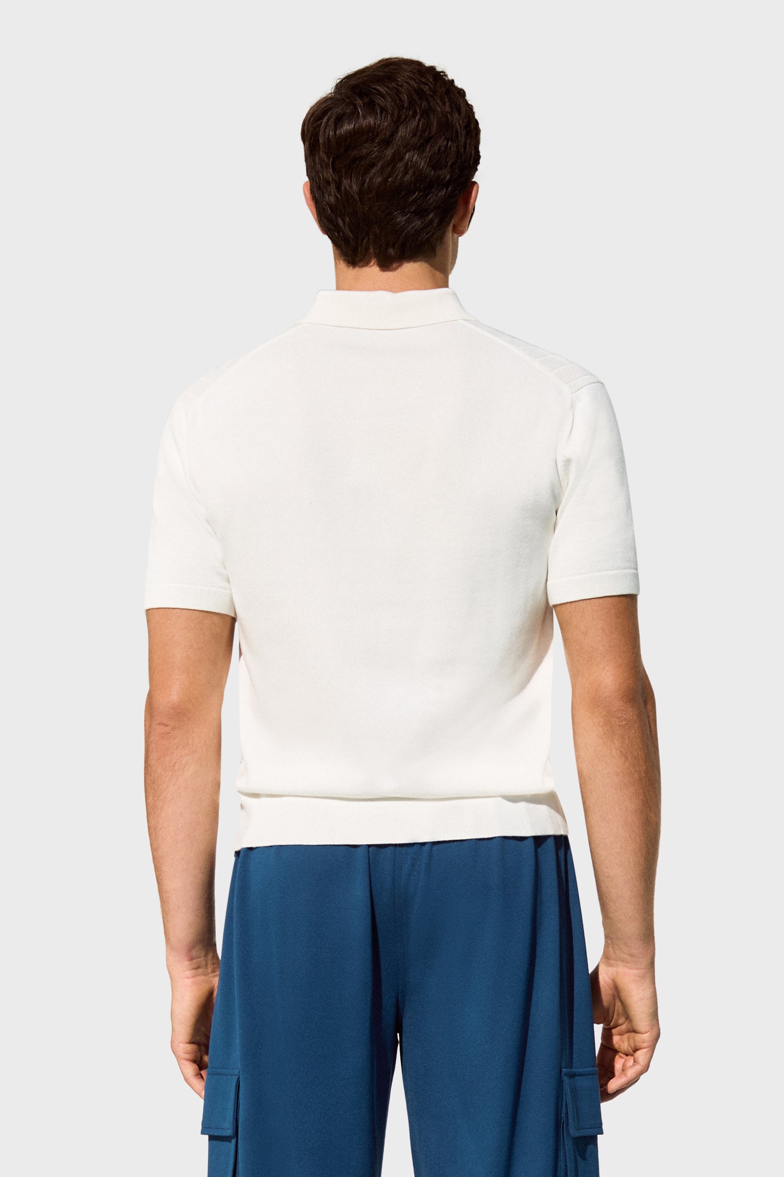 A man stands with his back to the camera, wearing the Alvarez Short-Sleeve Polo in white and blue cargo pants against a plain light gray background.