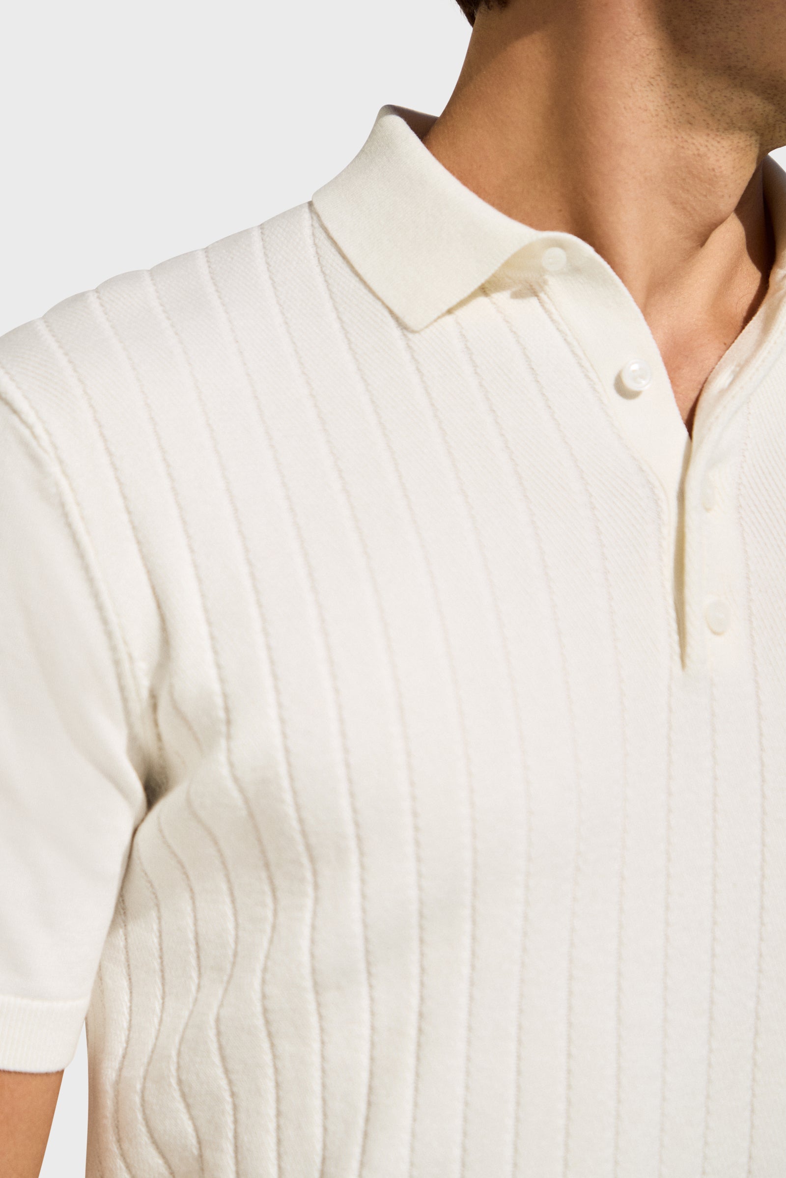 A close-up shows a person wearing the Alvarez Short-Sleeve Polo, featuring a white ribbed fabric, collar, and buttons. The image is cropped from shoulders to mid-chest against a plain, neutral background.