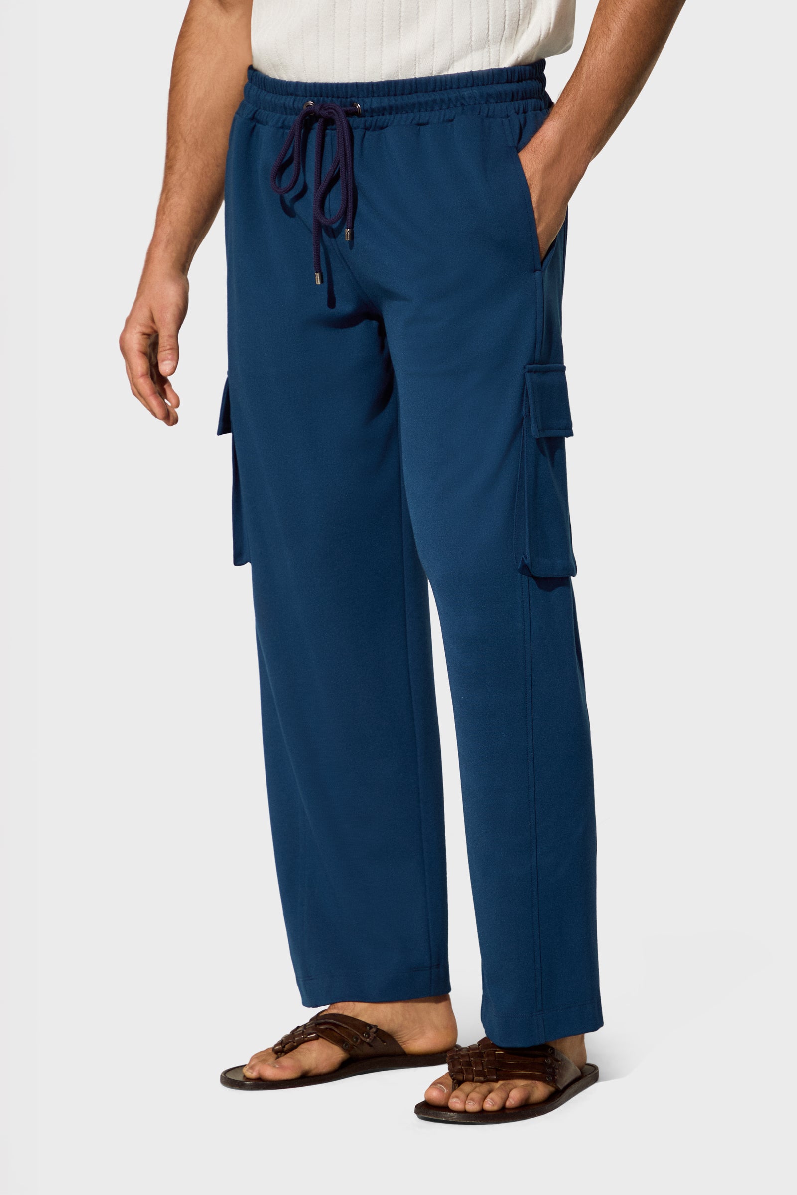 A person wearing blue Lounge Cargo Pants with side pockets, a white ribbed shirt, and brown sandals stands with hands in pockets against a plain light background.