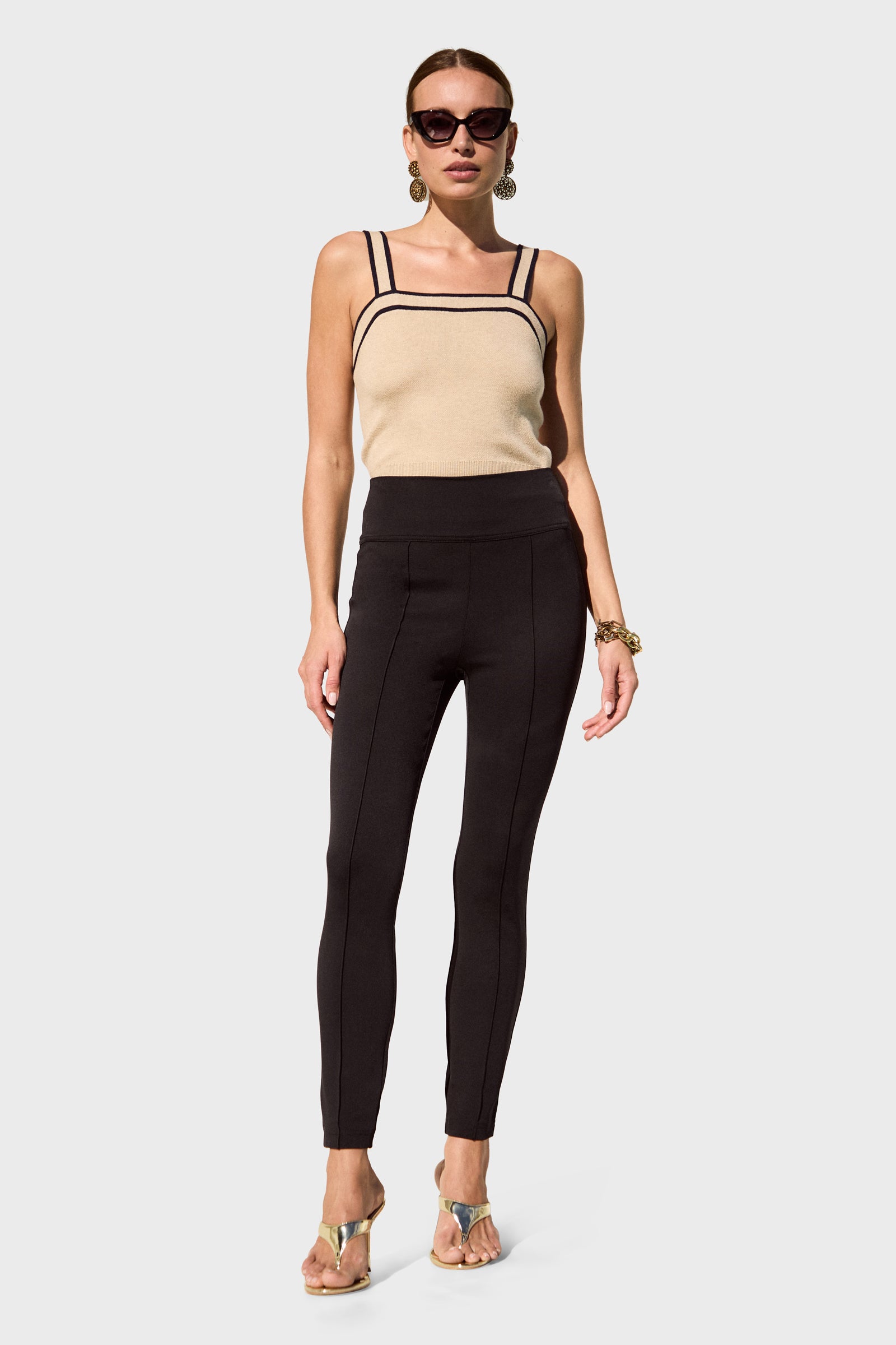 A woman poses against a plain background wearing sunglasses, a beige sleeveless top with black trim, Rose Slim-Leg Pants, heels, hoop earrings, and a bracelet.