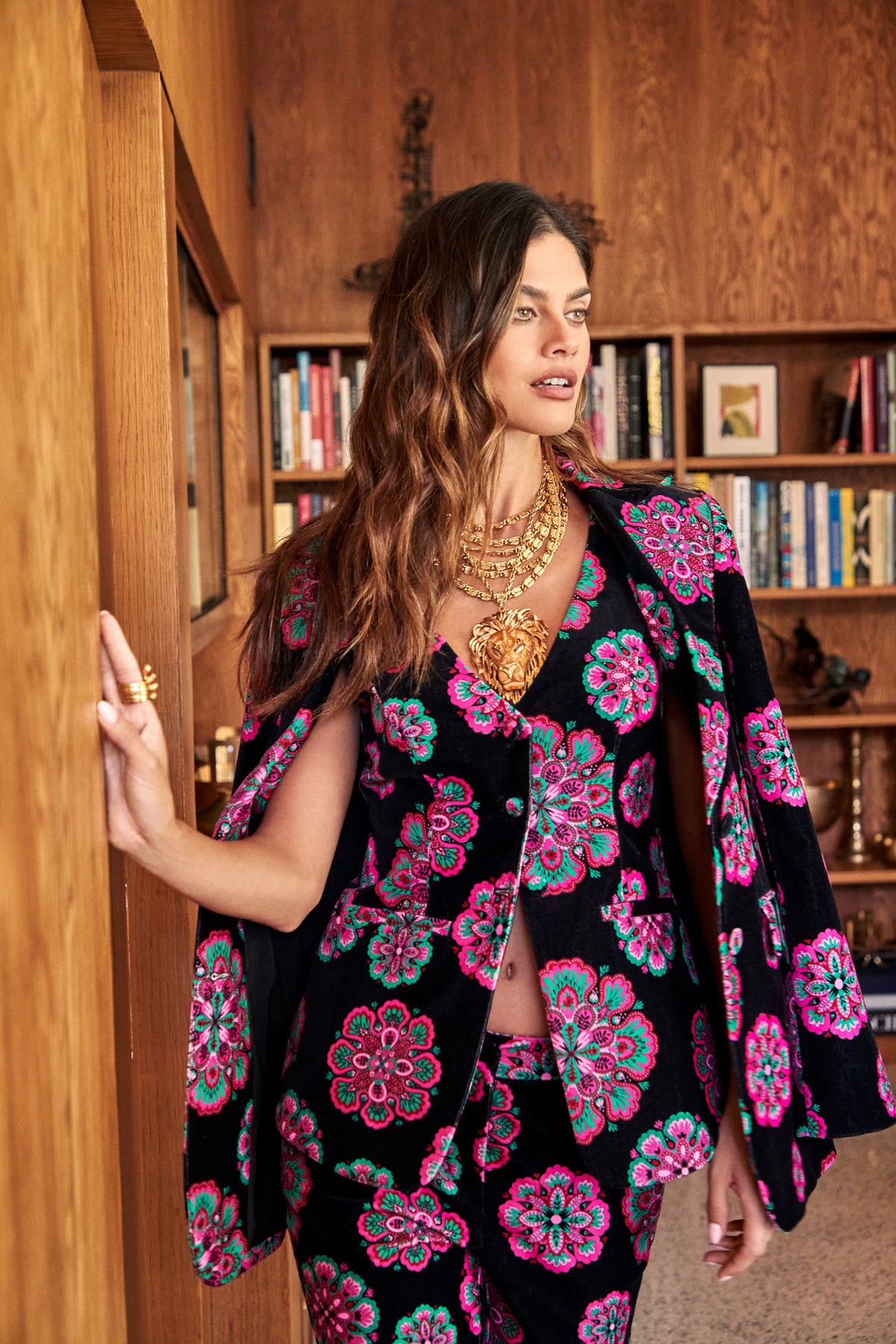 A woman wears a black, pink, and green floral patterned Raylee Top vest and matching blazer draped over her shoulders, styled with a layered gold statement necklace featuring a lion head pendant.
