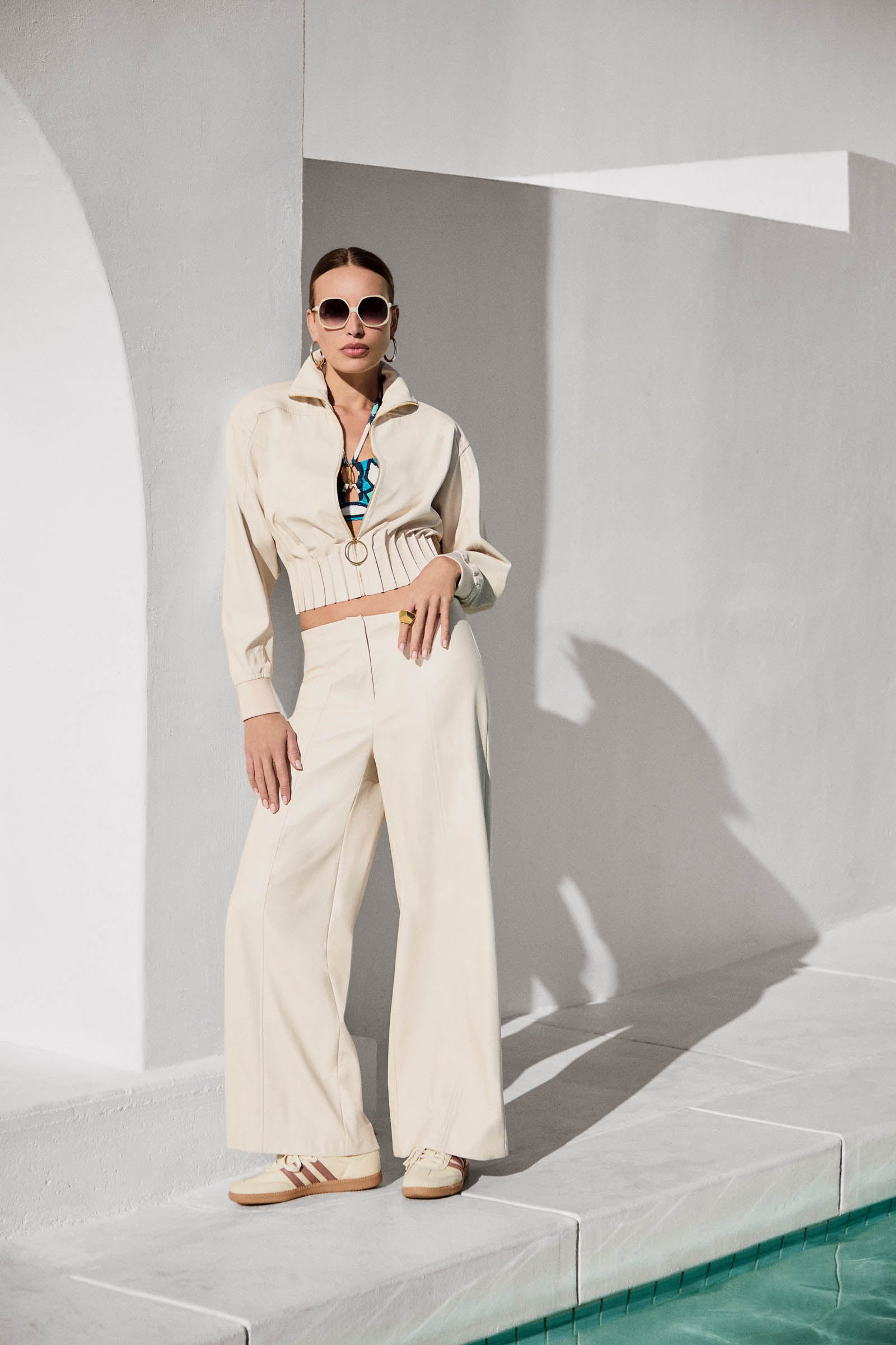 Wearing the Outrun Leatherette Jacket and cream wide-leg pants, a woman in sunglasses and sneakers stands confidently by a pool, posing against a white wall under bright sunlight with sparkling blue water beside her.