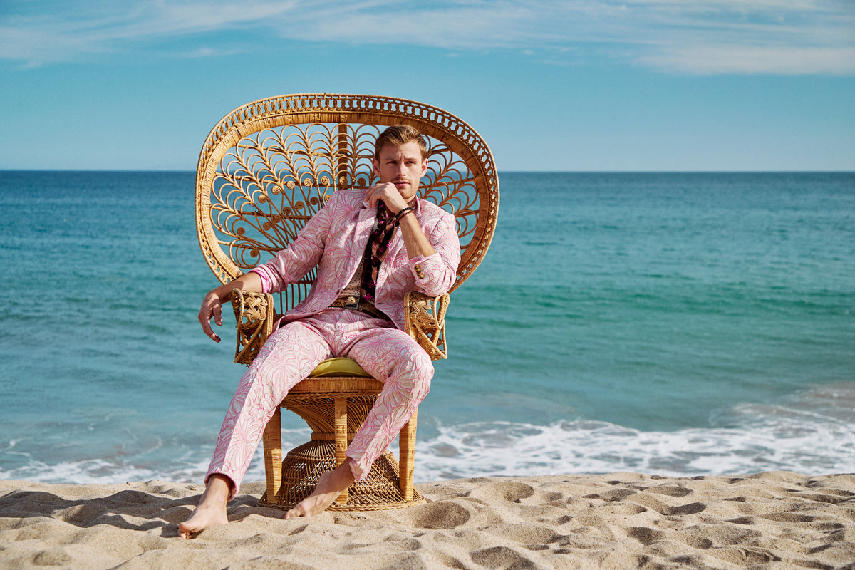 A man wearing the Thurston Blazer sits barefoot on a wicker chair on a sandy beach, with the blue ocean and sky in the background.