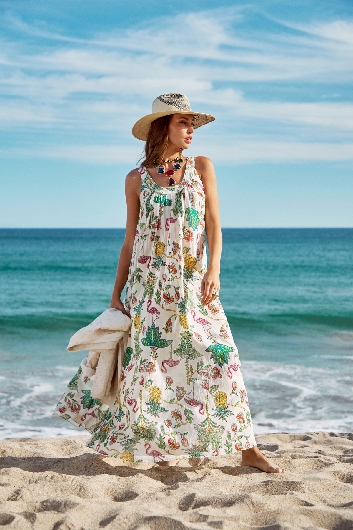 Wearing the Jianna Maxi Dress and a wide-brimmed hat, a woman stands on a sandy beach holding a towel, with the ocean and blue sky behind her.
