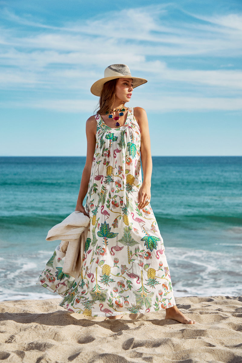 Wearing the Jianna Maxi Dress and a wide-brimmed hat, a woman stands on a sandy beach holding a towel, with the ocean and blue sky behind her.