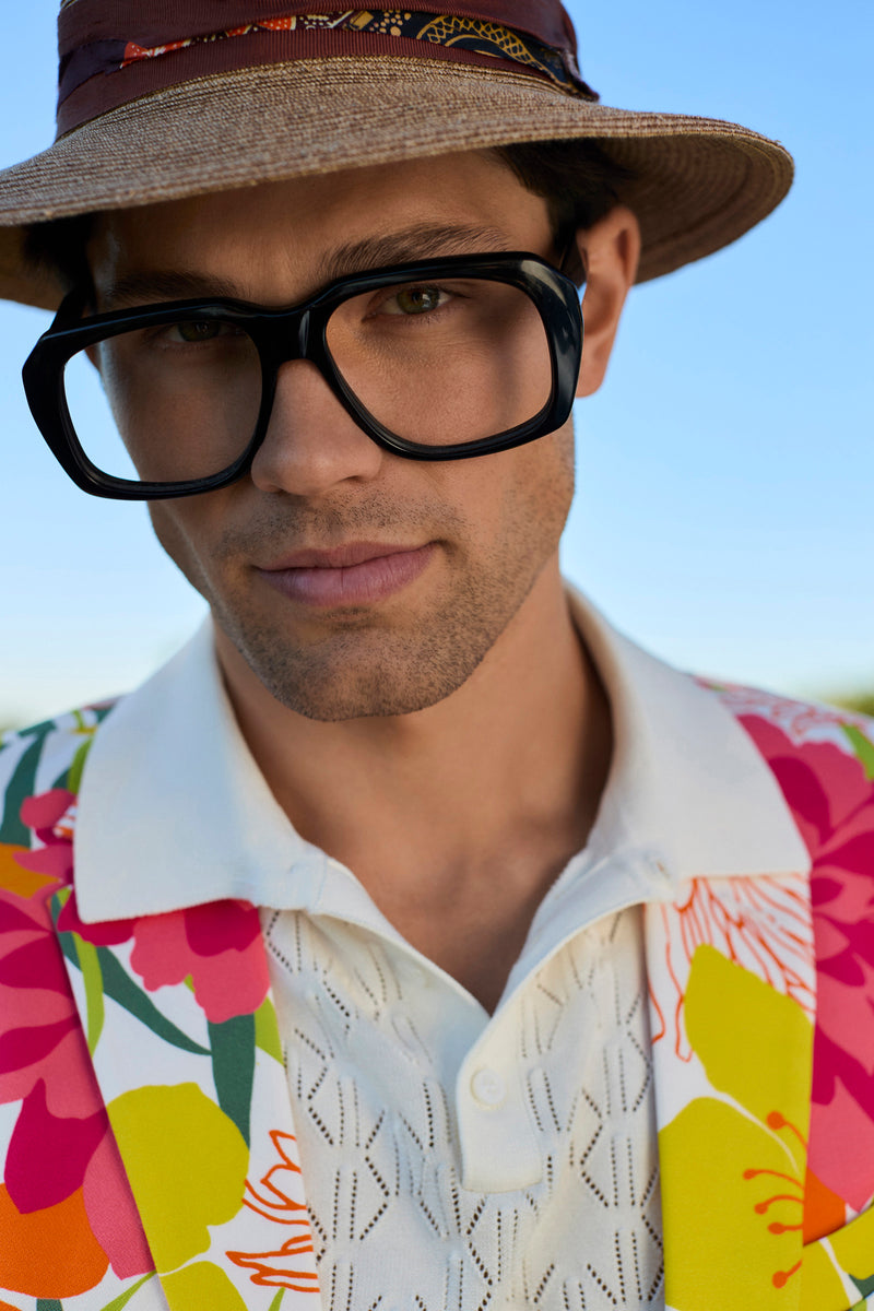 A person wearing the Luca Short-Sleeve Polo, a tan hat, large black glasses, and a colorful floral jacket stands outdoors under a blue sky.