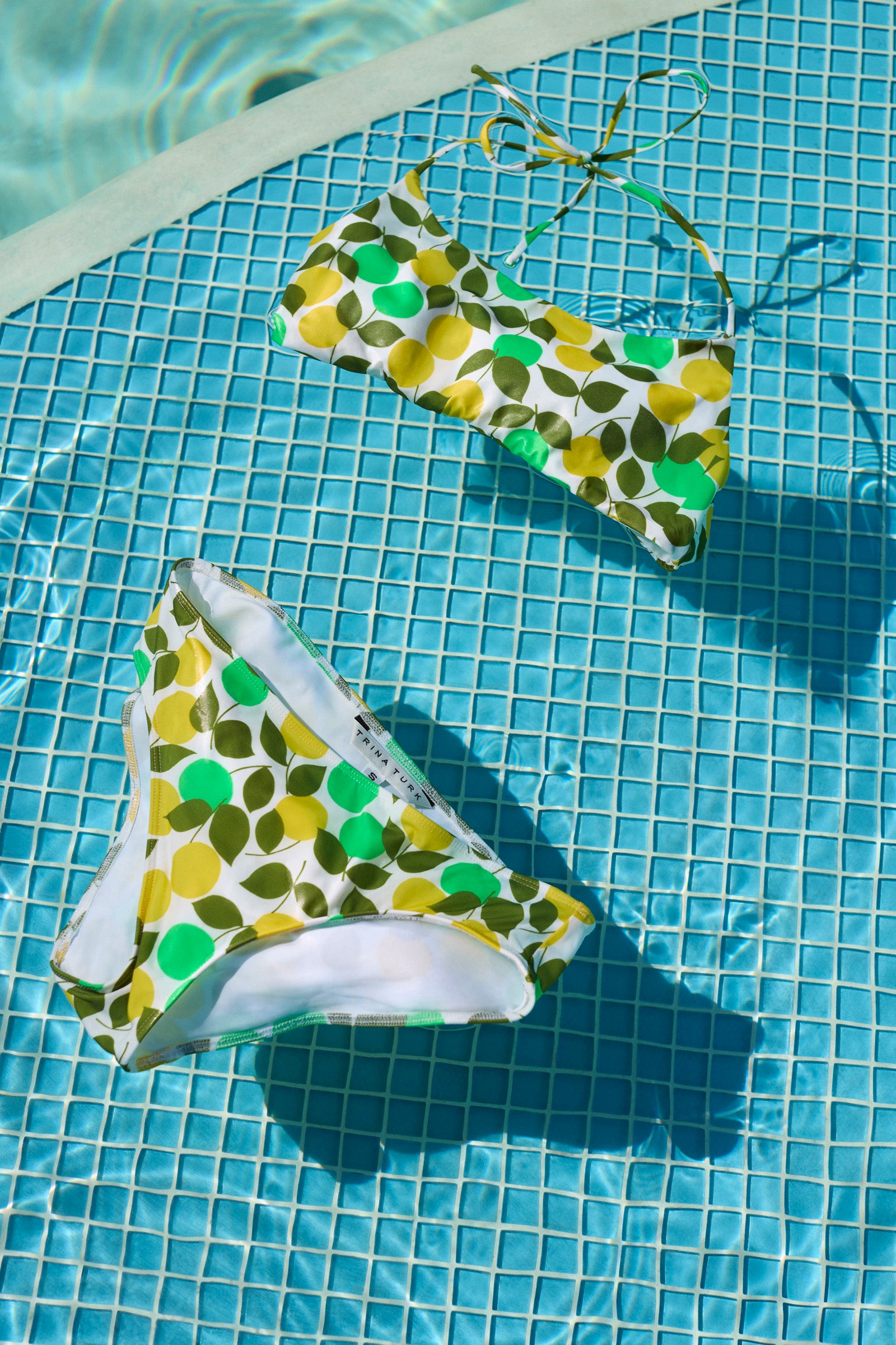 The Arissa Hipster Swim Bottom - Limone, with its green, yellow, and white leaf pattern, is displayed poolside on tiled flooring as sunlight casts shadows across the blue water tiles.