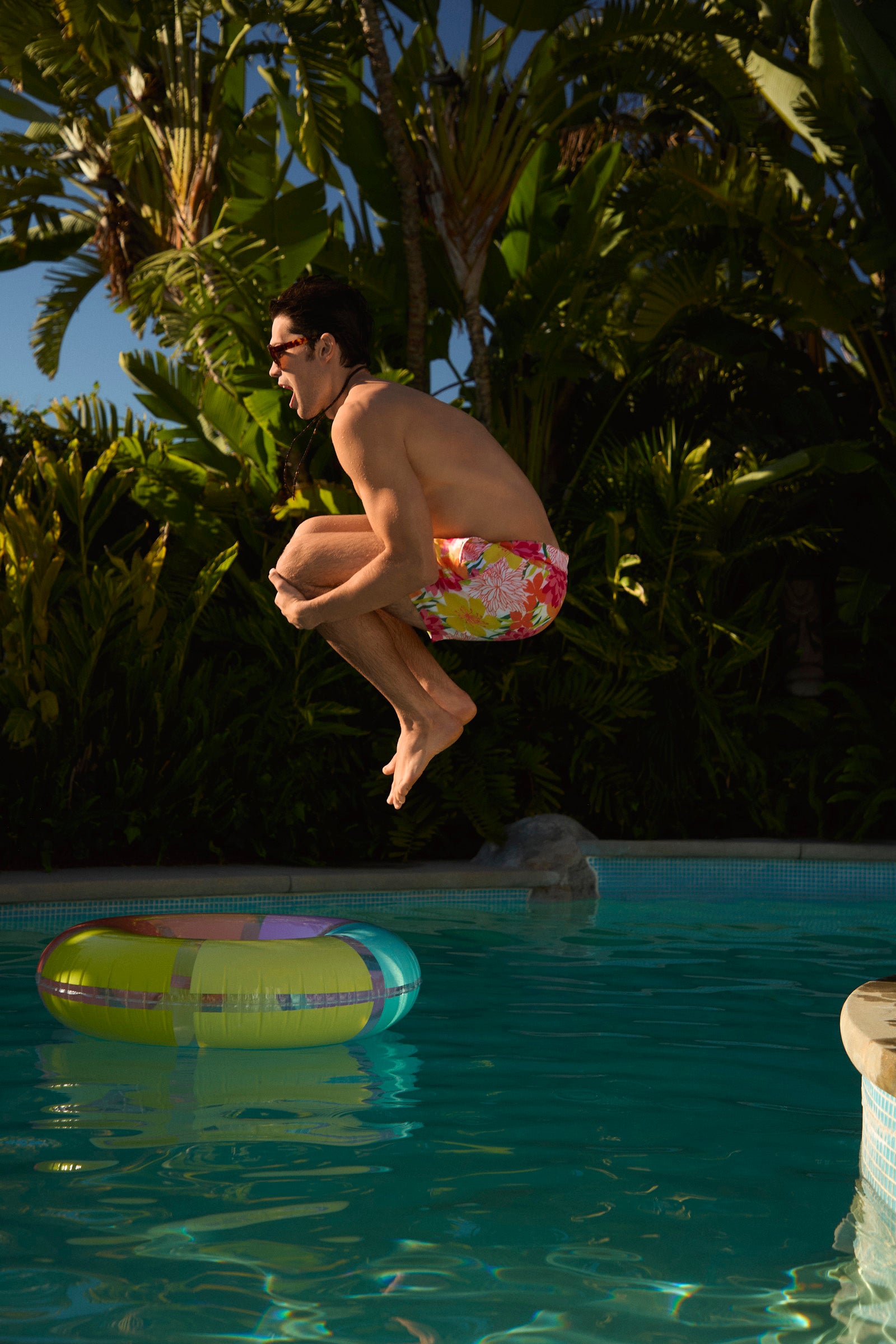 A man wearing Honolulu Board Short - Como leaps mid-air for a cannonball into a pool with a colorful inflatable ring, surrounded by tropical plants and palm trees.