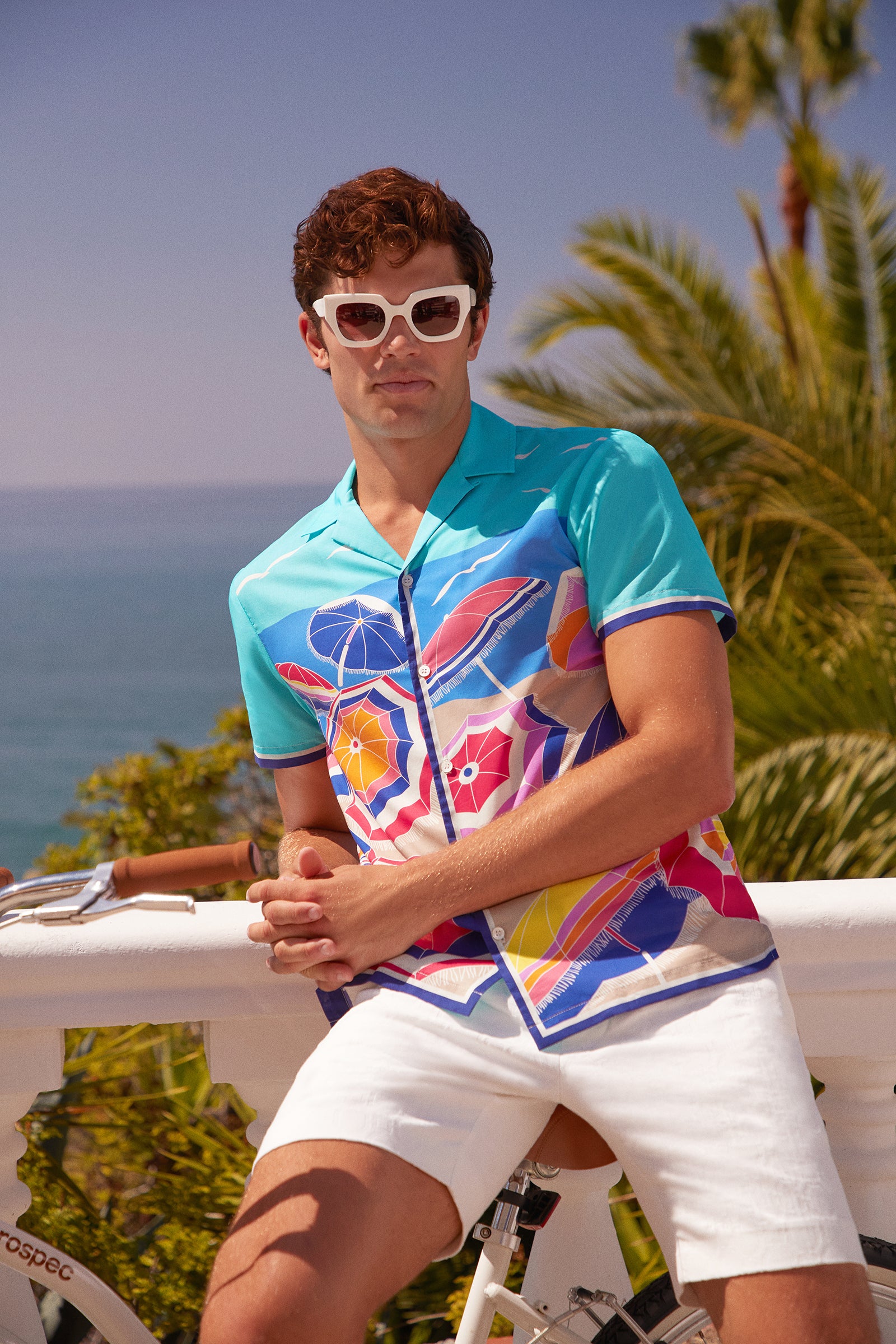A man in the Josue Short-Sleeve Shirt, white sunglasses, and white shorts leans against a white railing with palm trees and the ocean in the background.