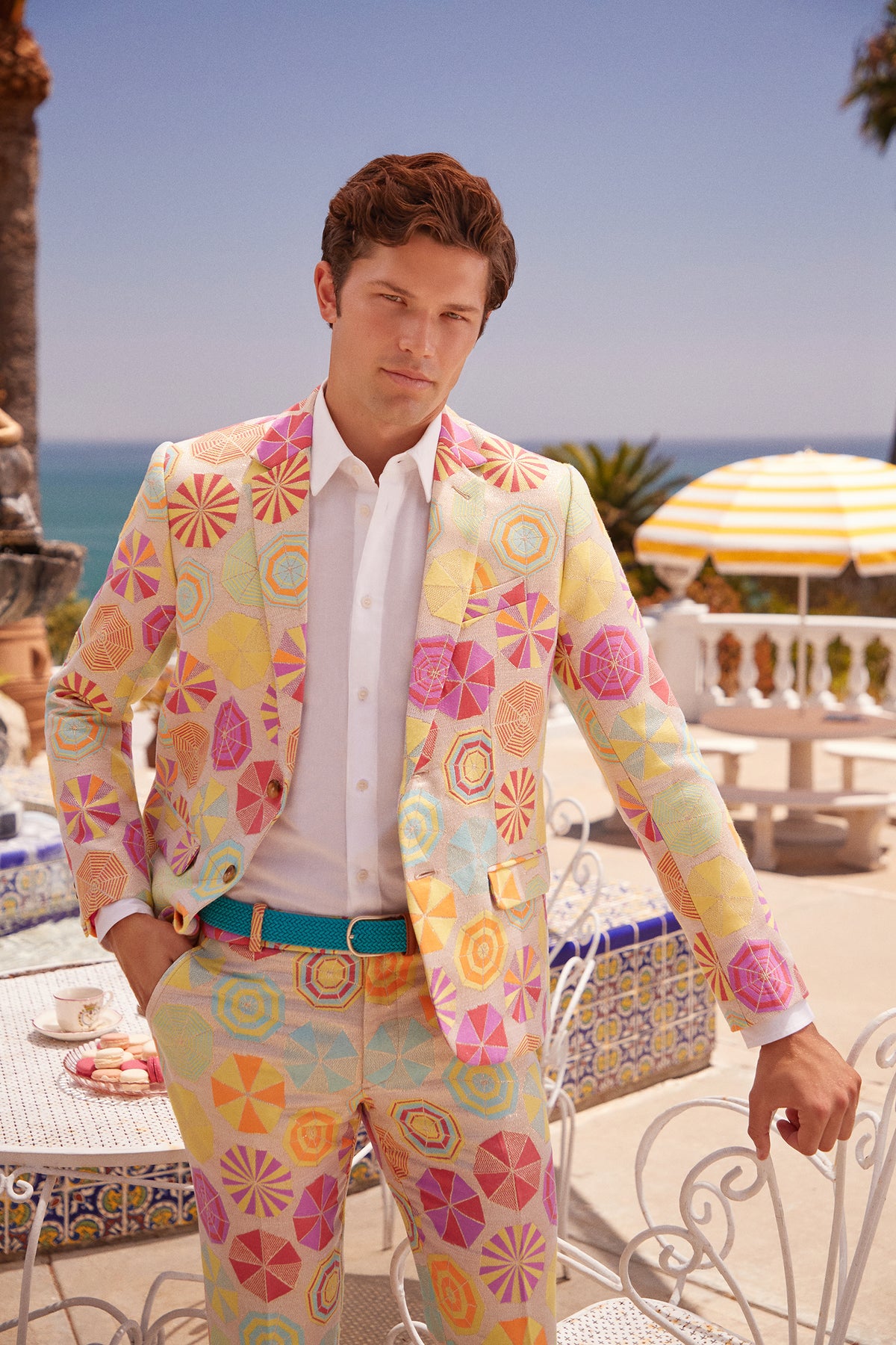 A man wearing the Thurston Blazer - Parapluie Jacquard stands outdoors by a white table and chairs, with the ocean, palm trees, and a yellow-striped umbrella in the background under a clear sky.