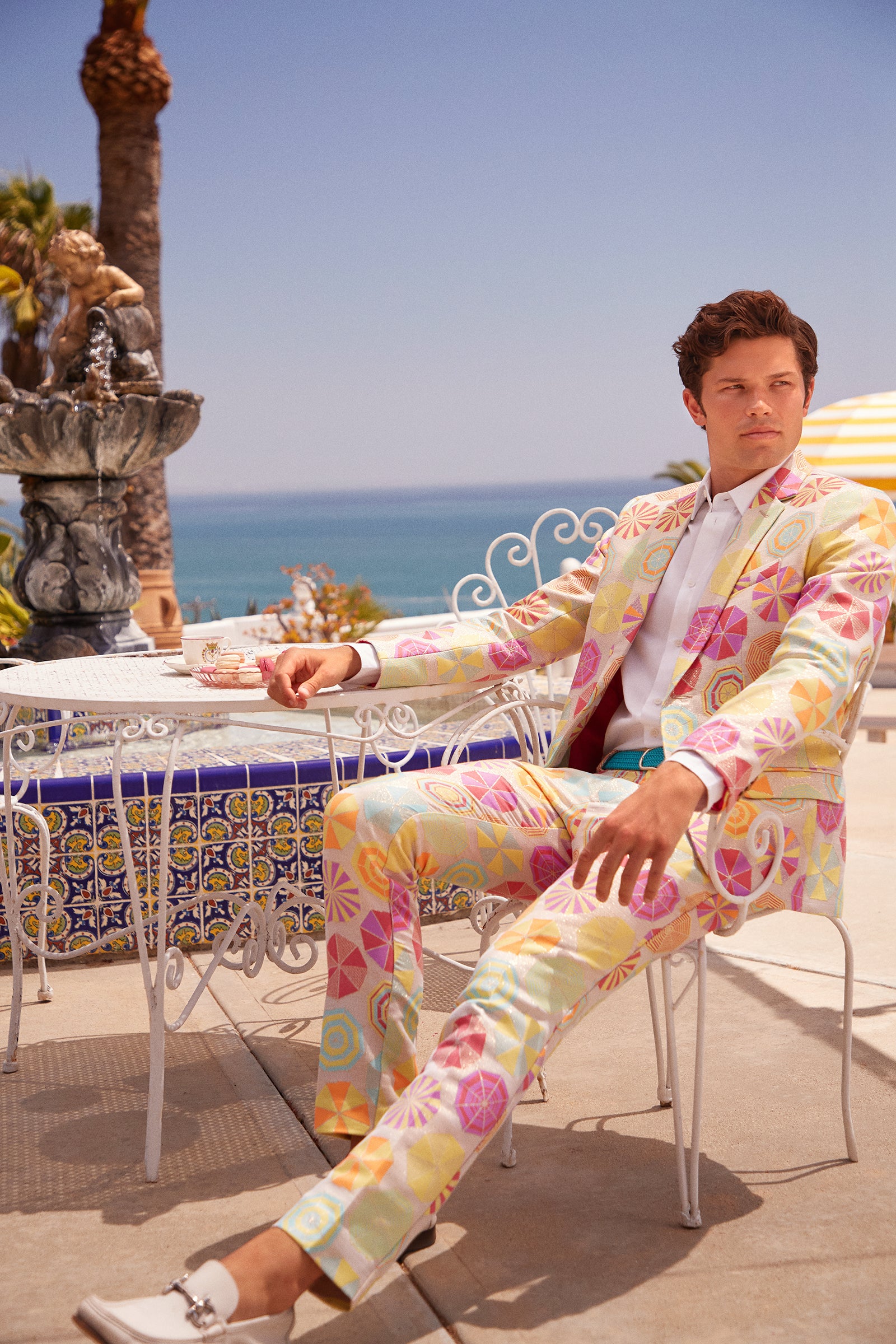 A man in the Clyde Slim Trouser - Parapluie Jacquard sits at a white wrought-iron table outdoors by a decorative fountain, with the sunny ocean and palm trees in the background.