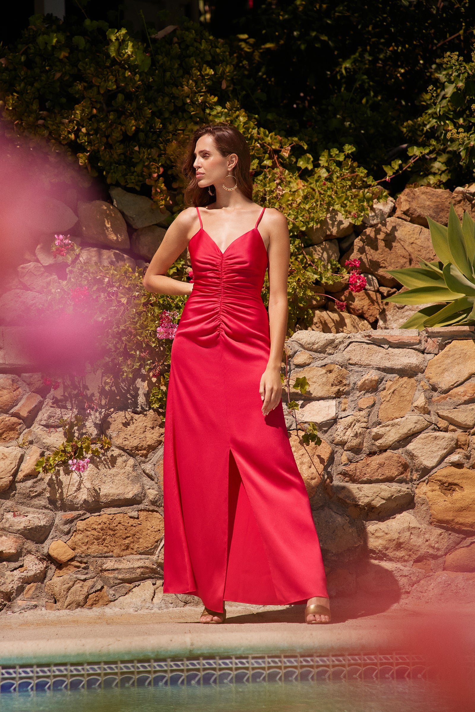 A woman wears the Tulipe Sleeveless Satin Dress in vibrant red, featuring a ruched front and thigh-high slit, standing by a stone wall with greenery and flowers near a pool under bright sunlight.