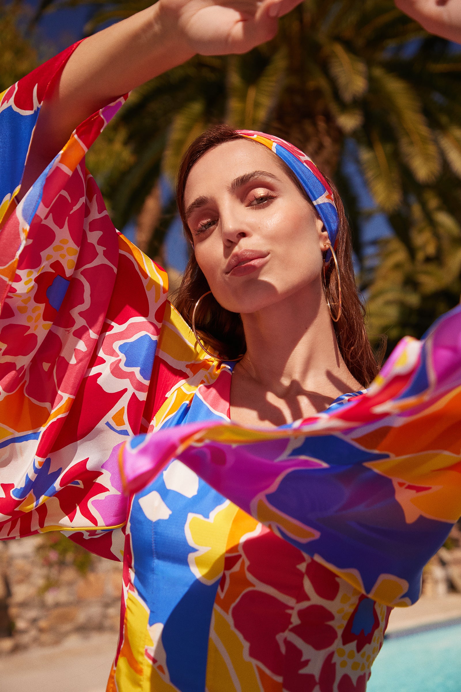 A woman in the Miera V-Neck Romper - Soie Paradis, hoop earrings, and a matching headband poses with raised arms under palm trees in bright tropical sunlight.
