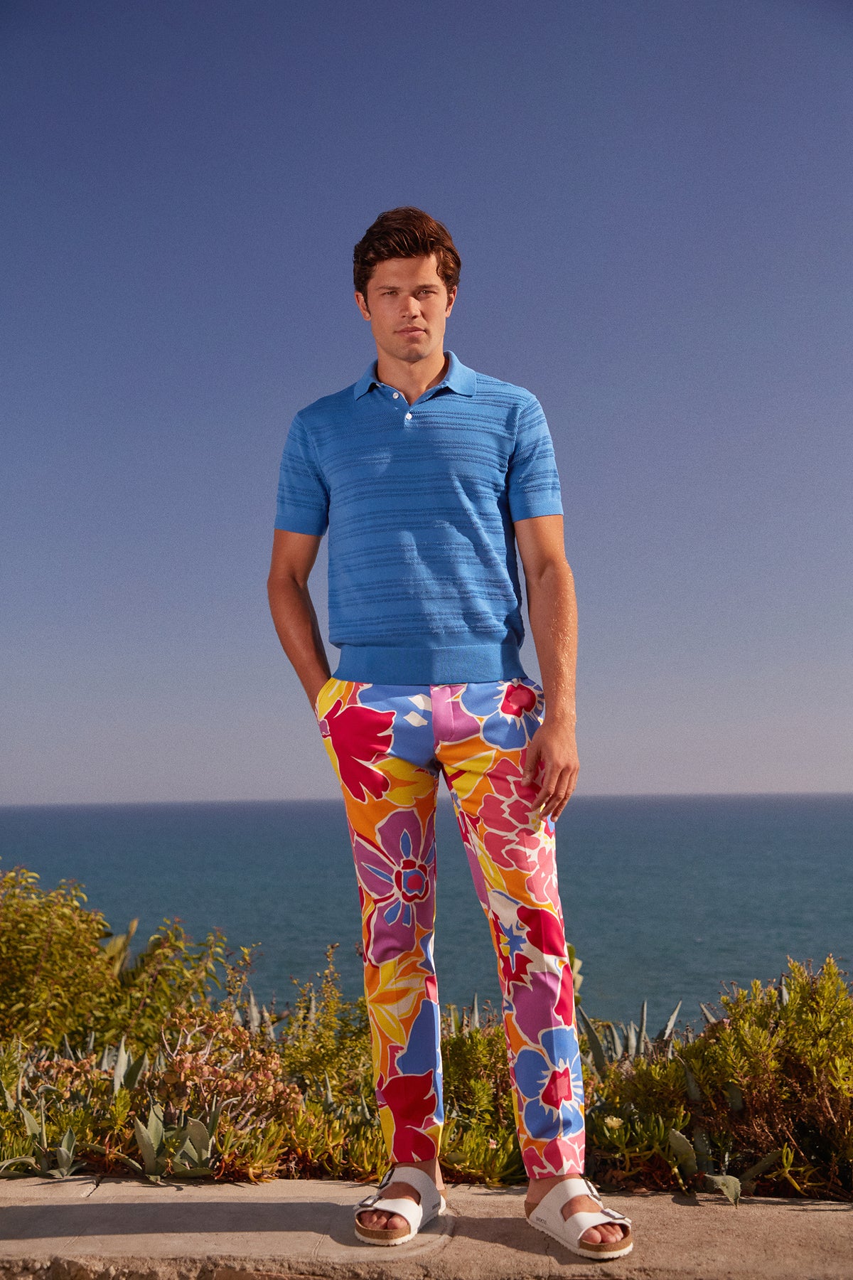 A man stands by the ocean in the Clyde Slim Trouser - Soie Paradis, featuring colorful floral prints, paired with a light blue polo shirt and white sandals. The clear blue sky, greenery, and water create a serene backdrop.